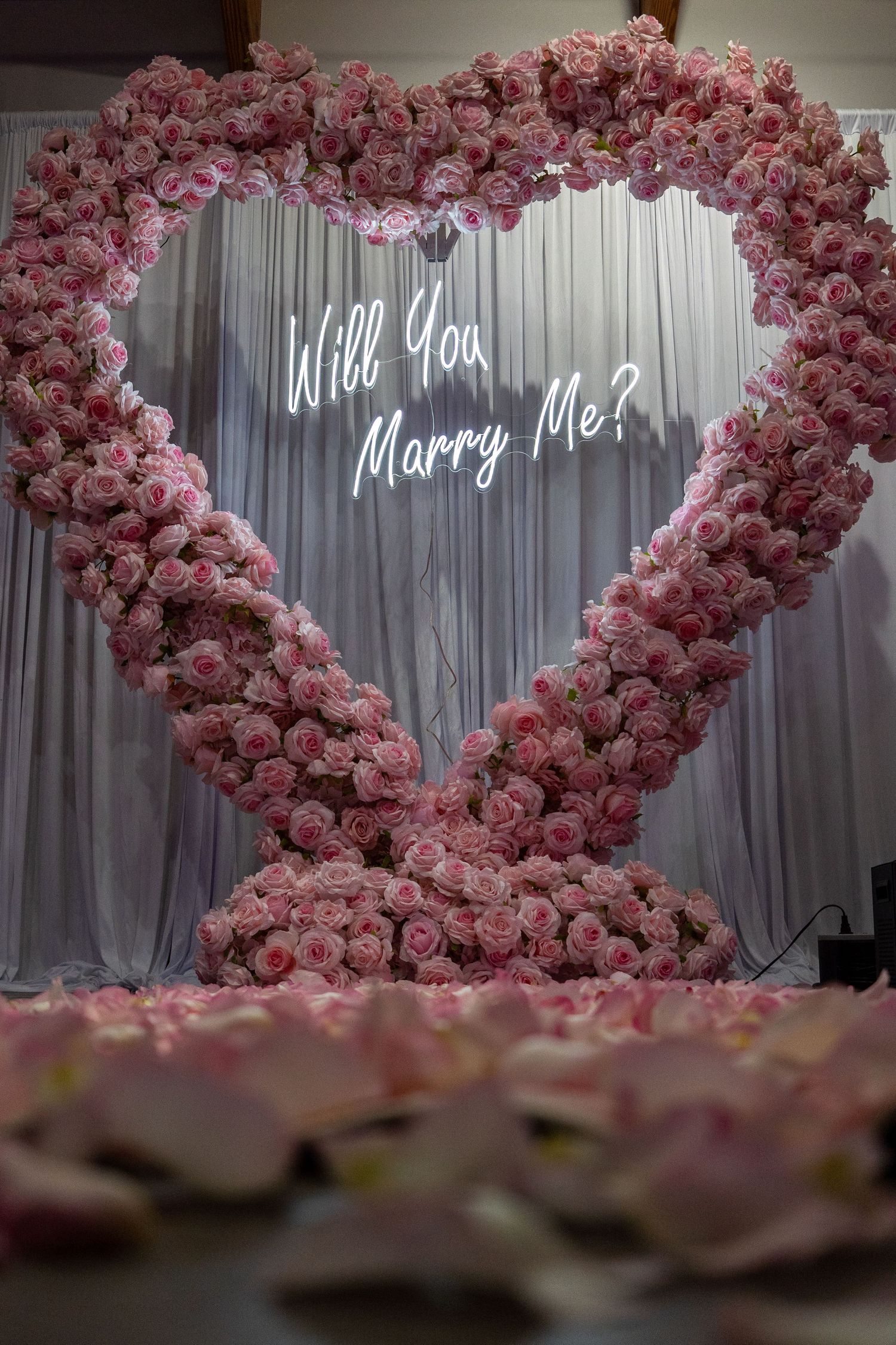 Pink floral arch with illuminated 'Marry Me?' sign creates romantic proposal setting.