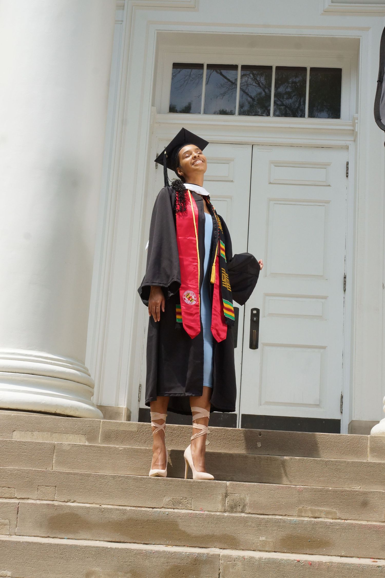 Graduate in black cap and gown with colorful stoles poses proudly on steps before white columned building.