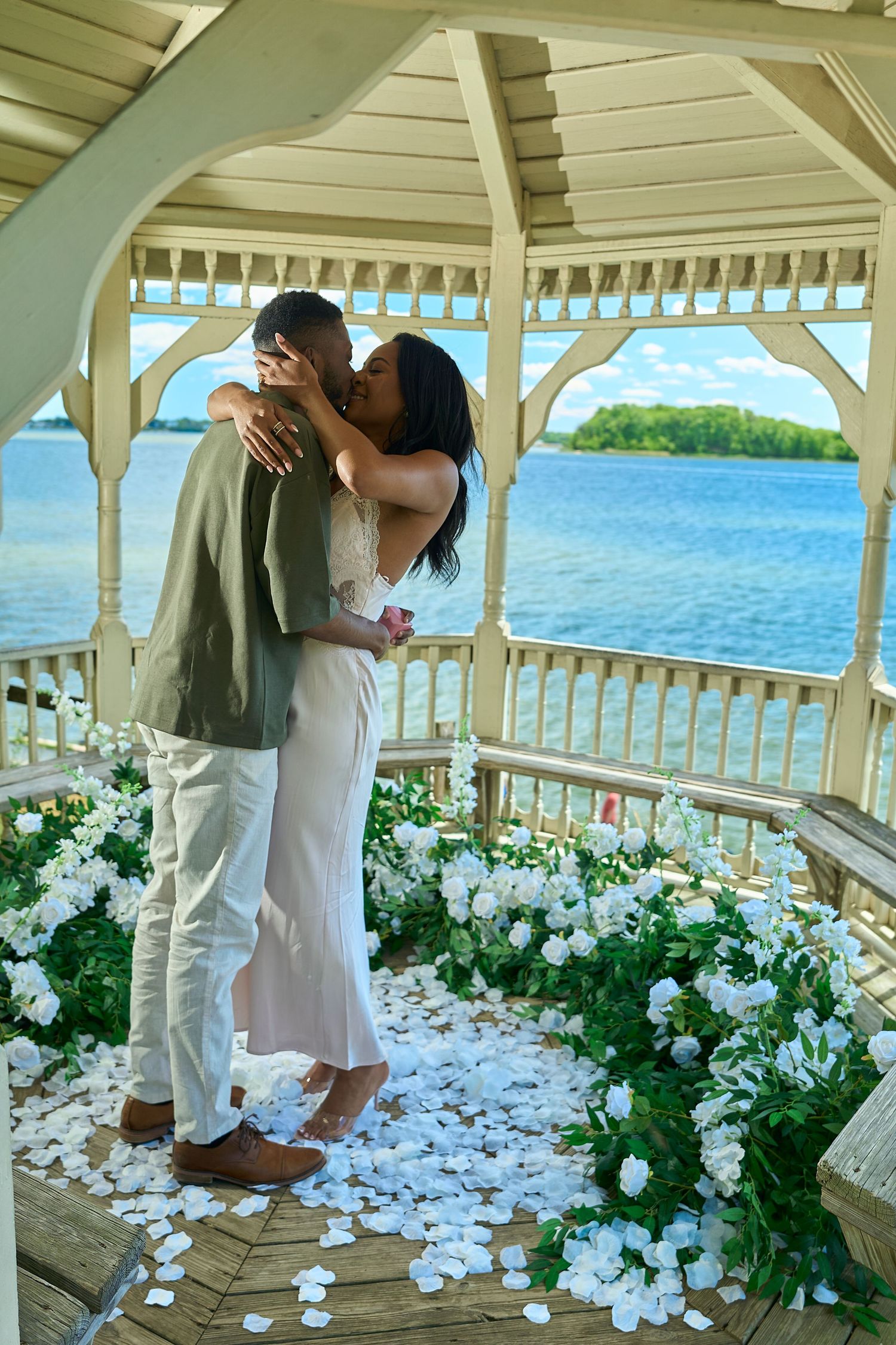 Couple sharing a romantic kiss in a white gazebo decorated with white flowers and rose petals overlooking a serene blue waterway.