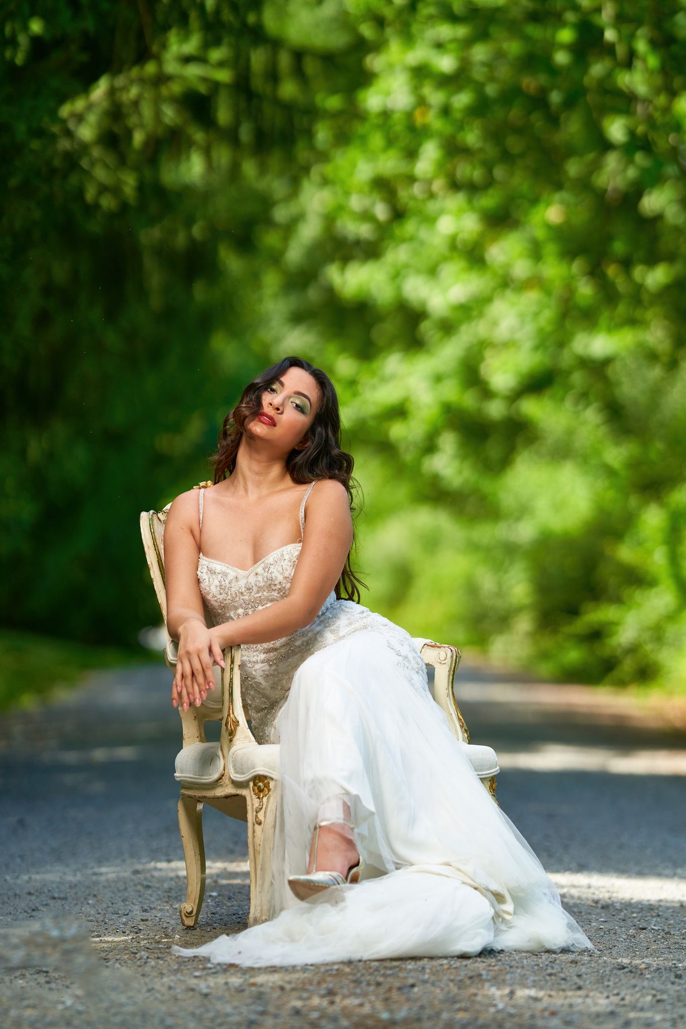 A bride in a white wedding dress poses elegantly on a vintage chair outdoors with a lush green forest backdrop.