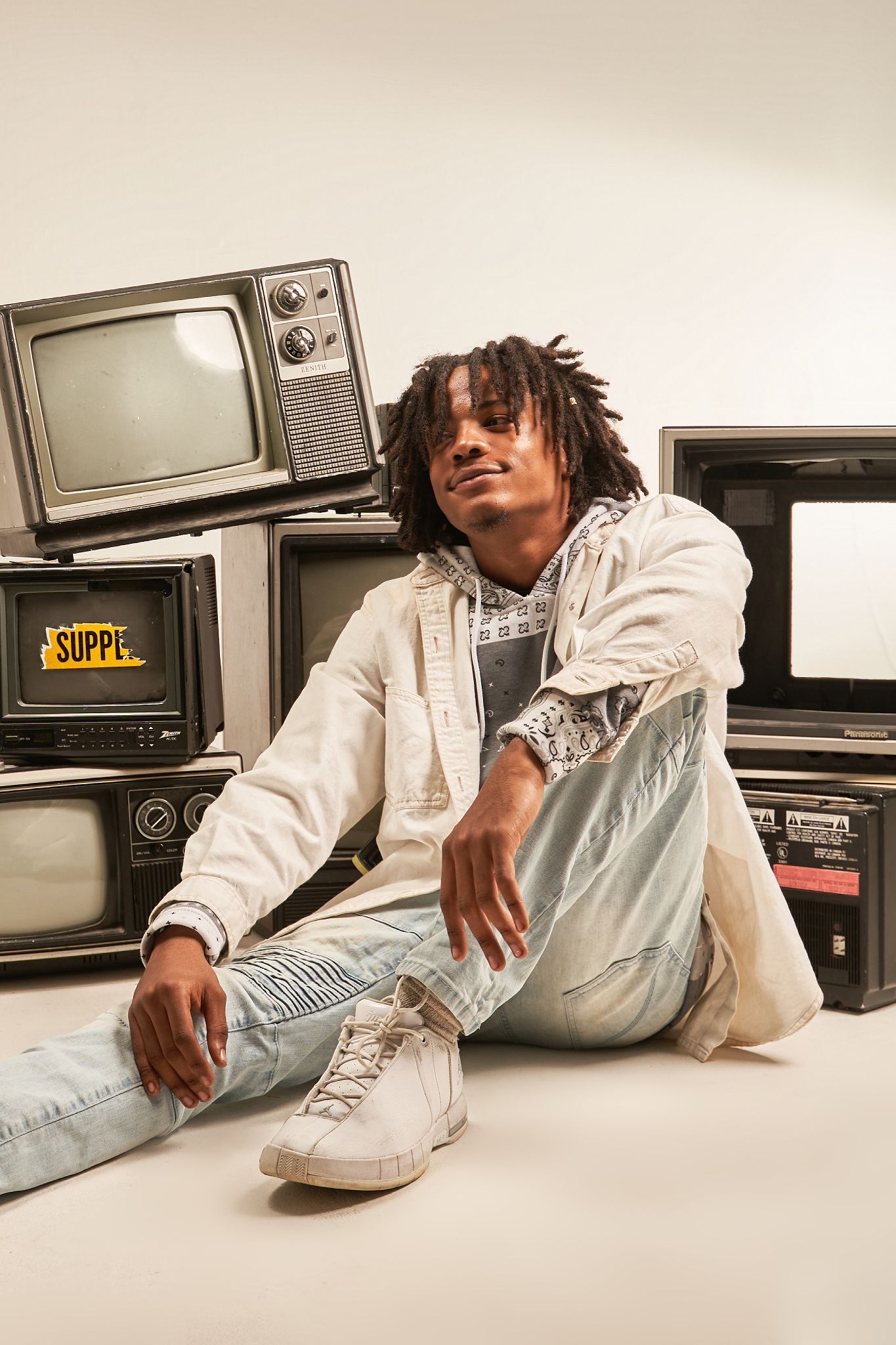 Person in white outfit lounging on floor surrounded by vintage television sets.