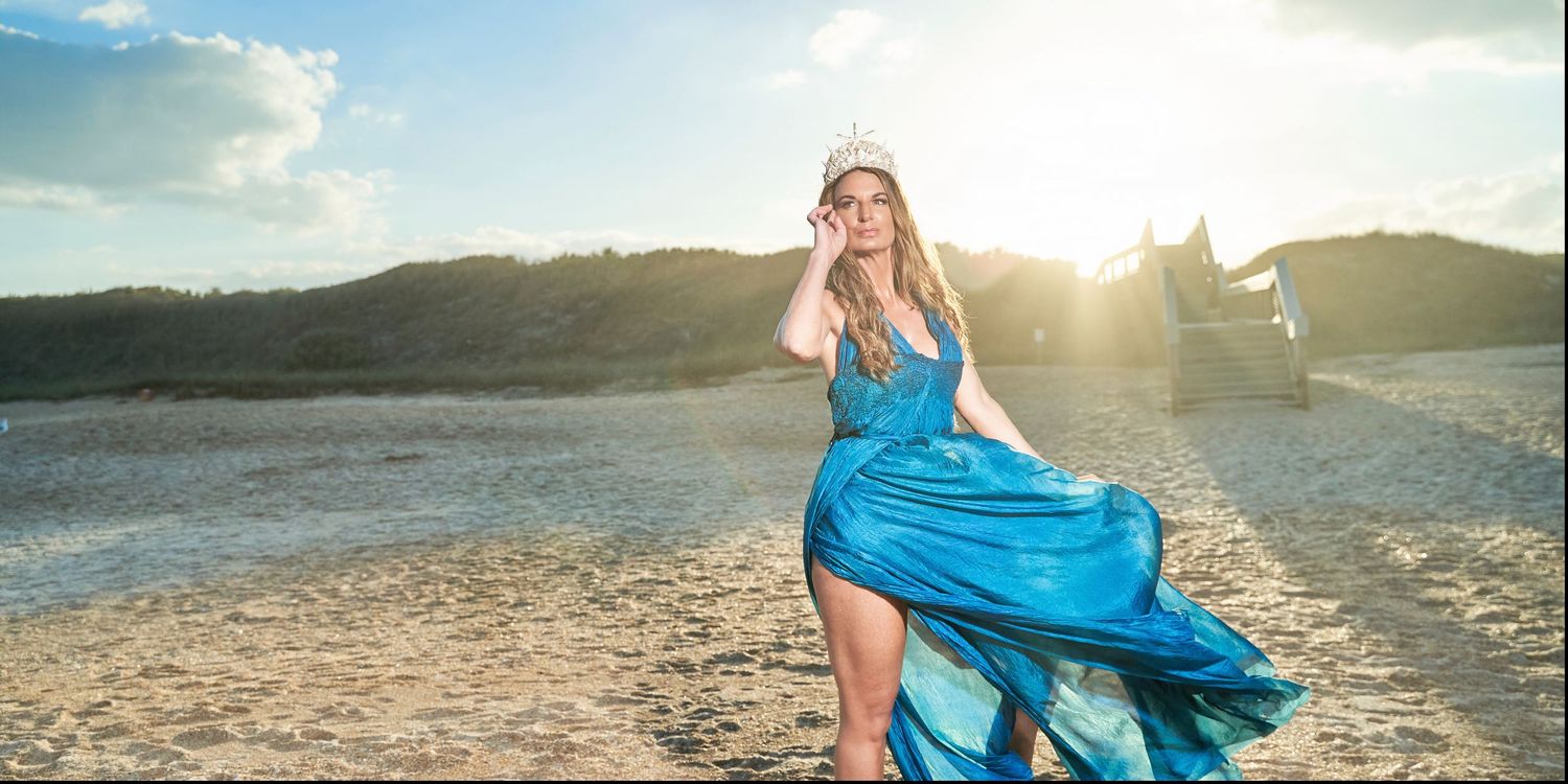 Woman in flowing blue dress poses dramatically on beach at sunset.