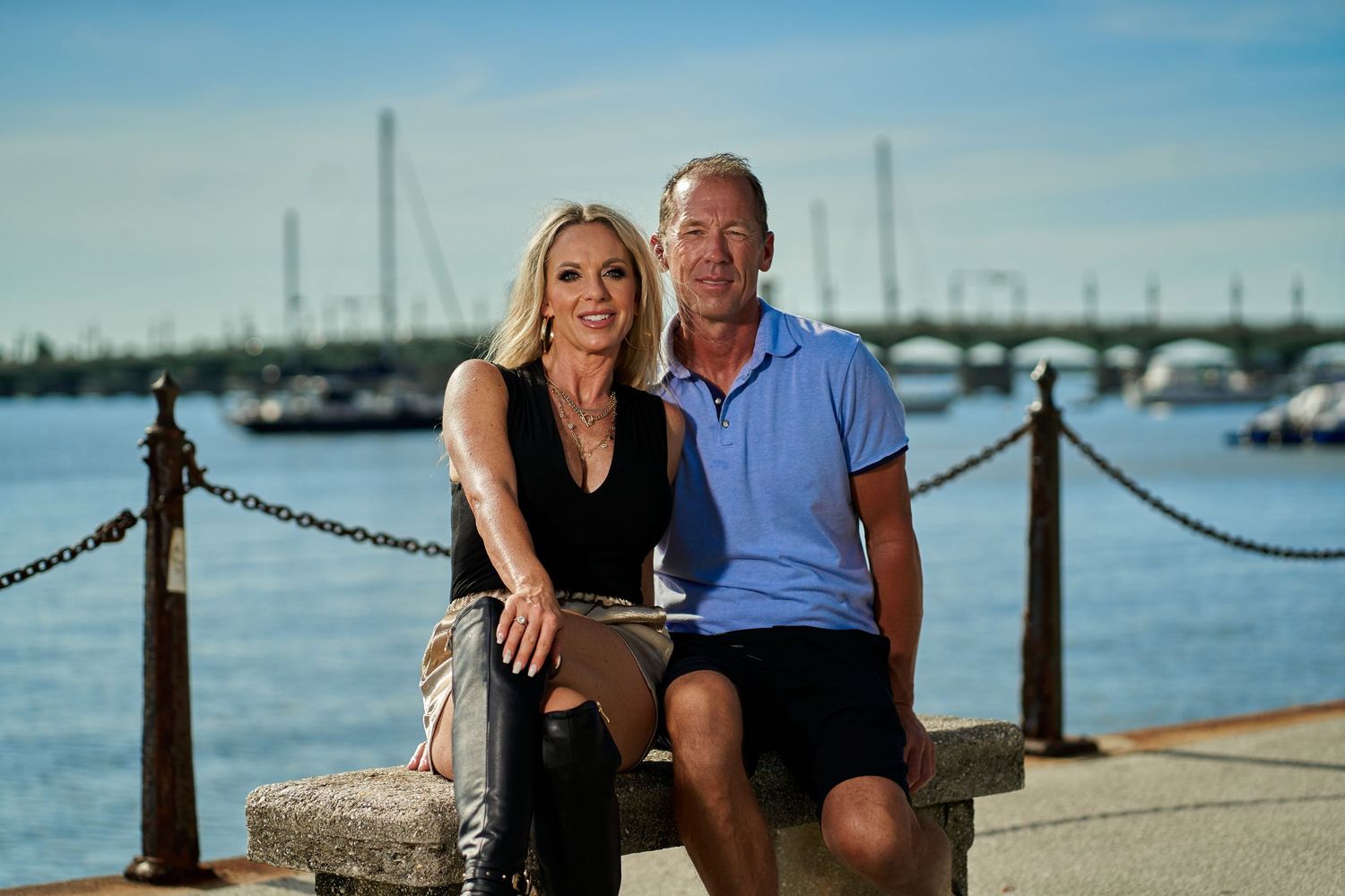 Couple sitting together on a waterfront pier with boats and marina in the background.