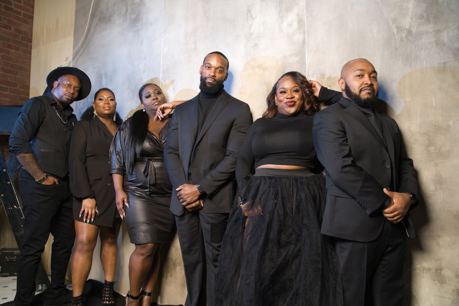Group wearing formal black attire poses together against a textured wall.