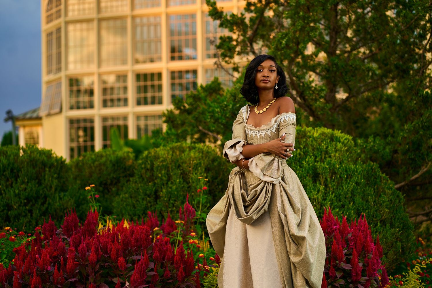 Elegant period dress photographed in front of historic stone building with red flowers in foreground.