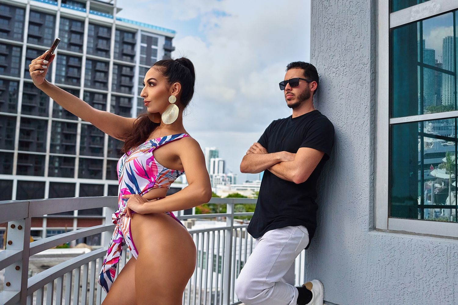 Two people taking selfies on a sunny balcony overlooking city buildings.