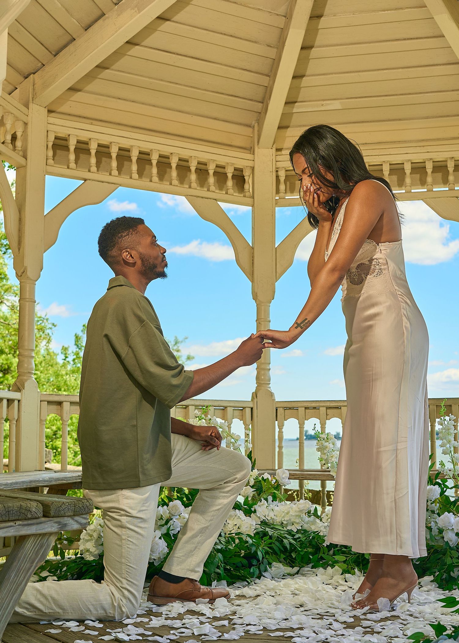 Man proposes on one knee to woman in a white floral gazebo with blue sky backdrop.