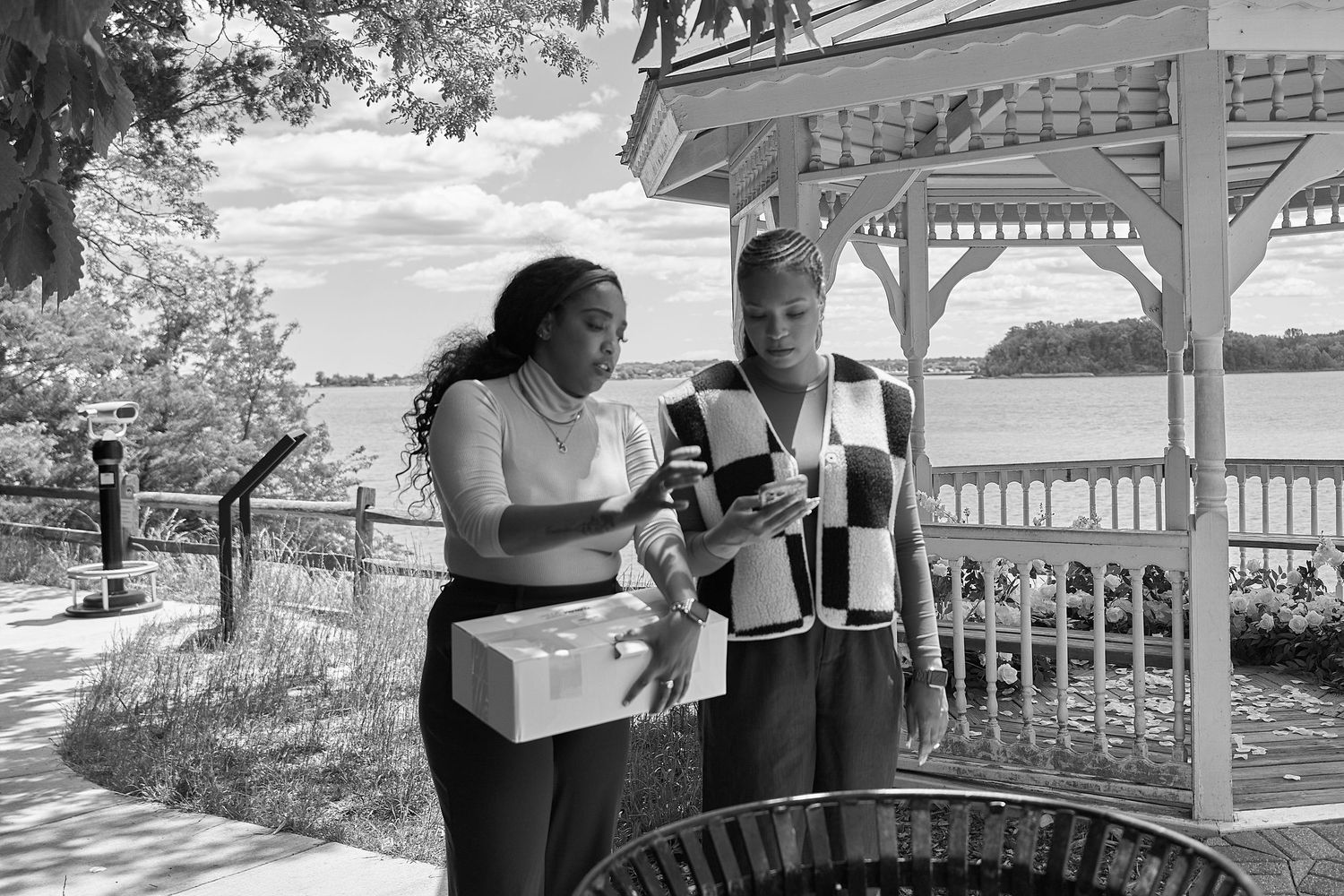 Two women stand near a gazebo by a lake, one holding a box, in a black and white outdoor photo.