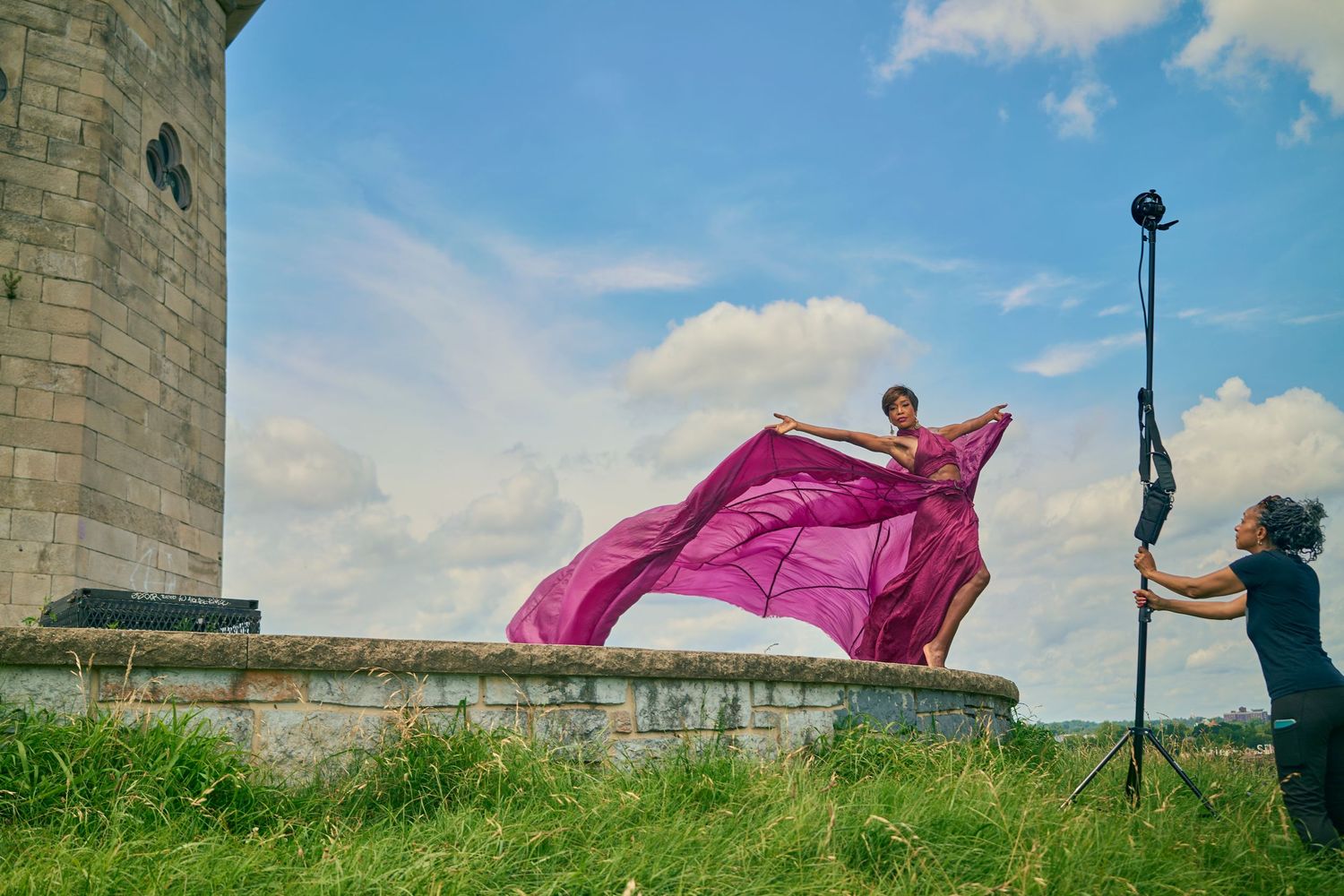 Behind-the-scenes fashion shoot with model in pink flowing gown near stone tower with photographer assistant.