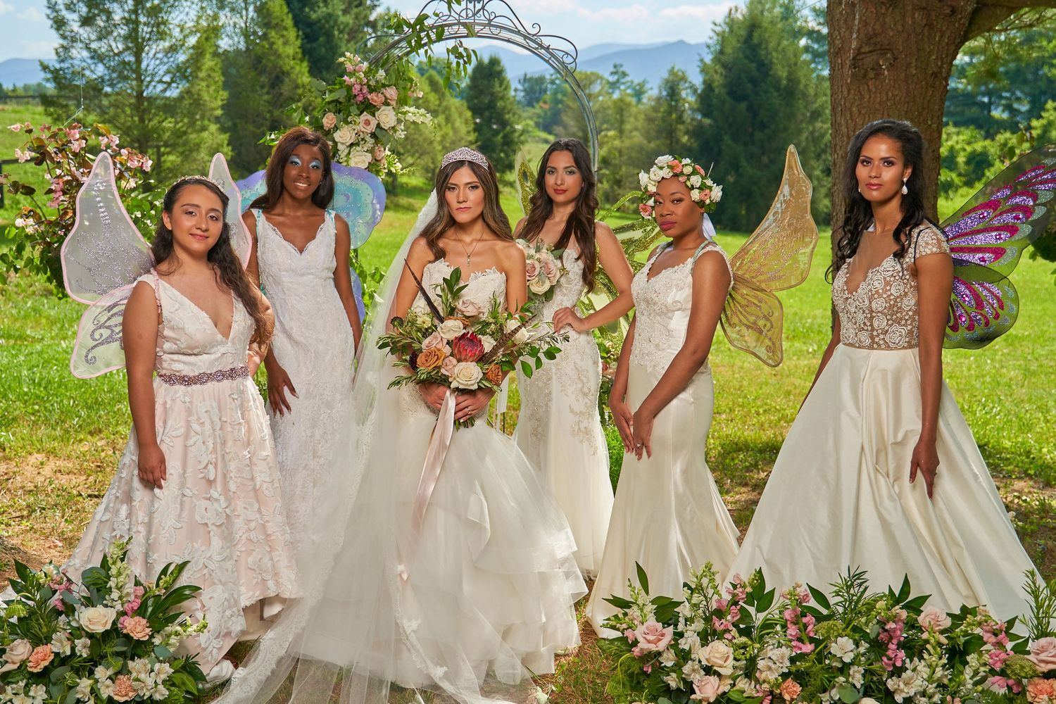 Wedding party in white dresses poses outdoors with mountain scenery in background.