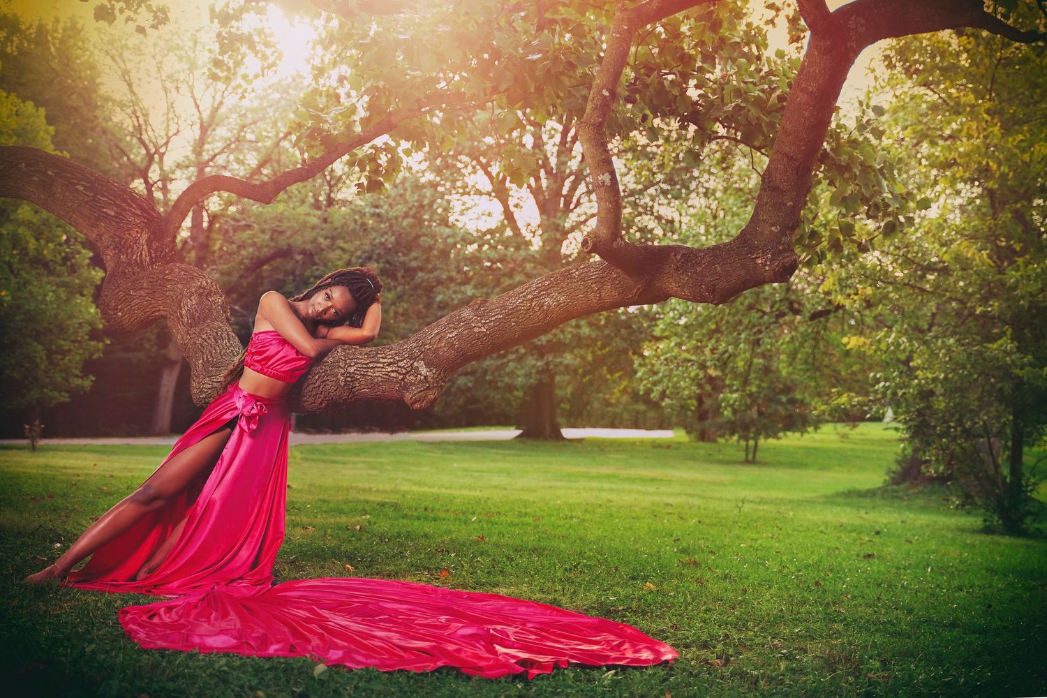 Dramatic red dress flows in breeze as model poses beneath sunlit tree in park setting.