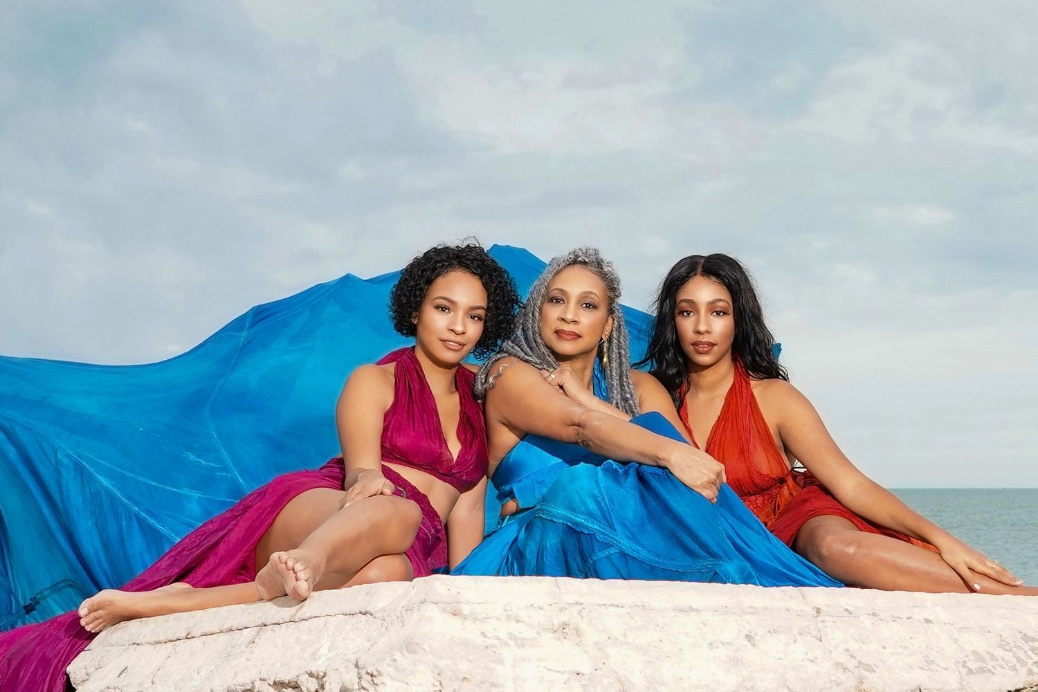 Group of women in colorful swimwear posing together on a beach with blue fabric and sand.