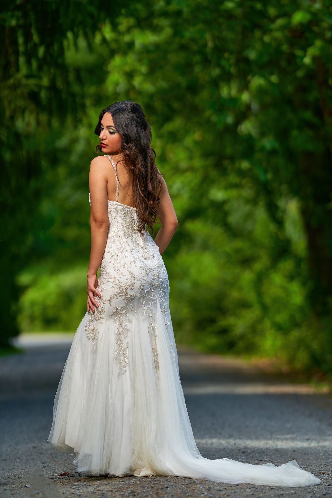 A bride in a flowing white lace wedding gown with tulle skirt poses on a tree-lined road during a photo session.
