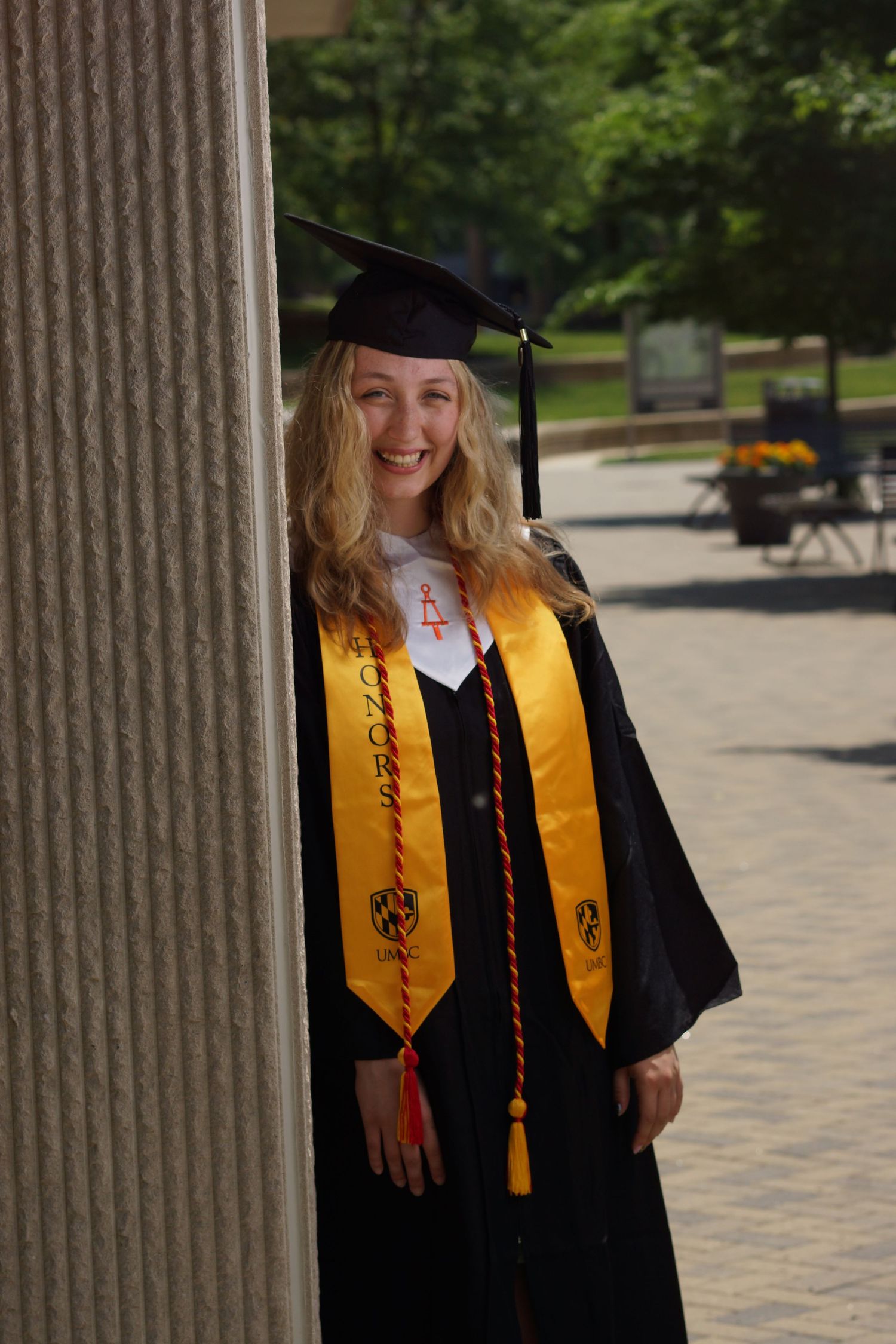 Smiling female graduate in black cap and gown with gold honor stole poses beside a column on campus.
