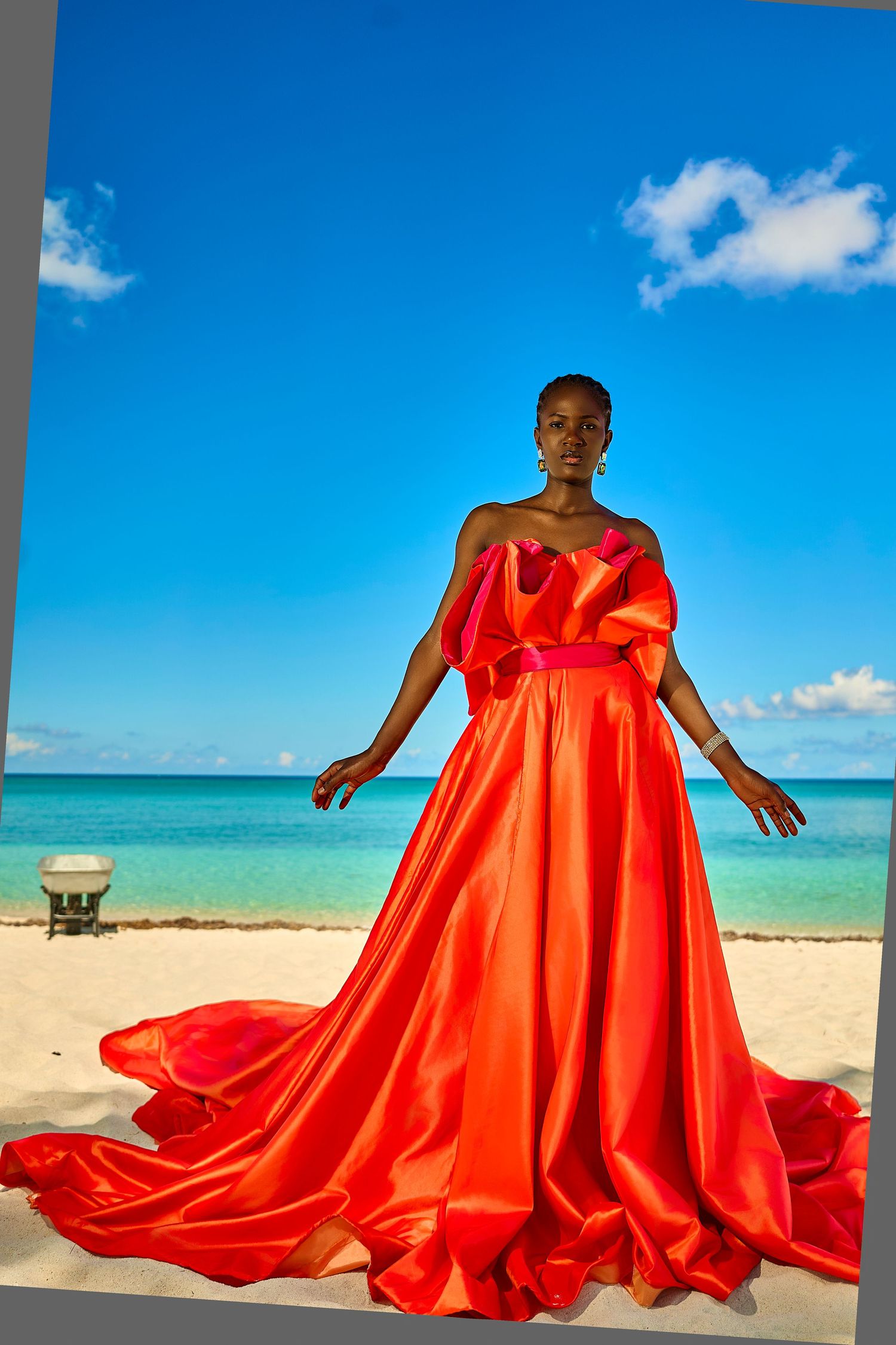 Woman in flowing red satin gown poses on tropical beach with turquoise water and blue sky.