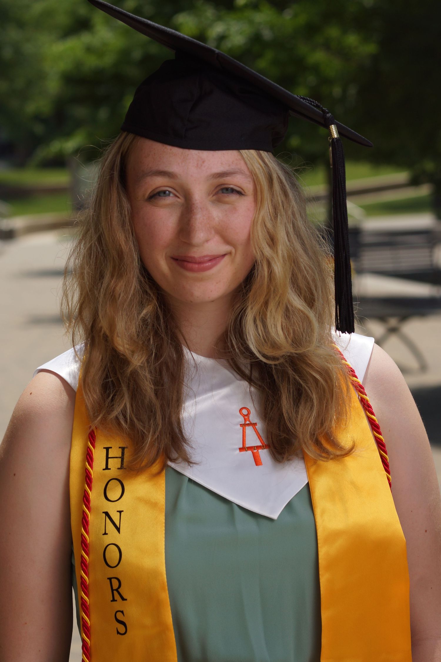 Smiling female graduate wearing black cap, honors sash, and white collar at outdoor graduation ceremony.