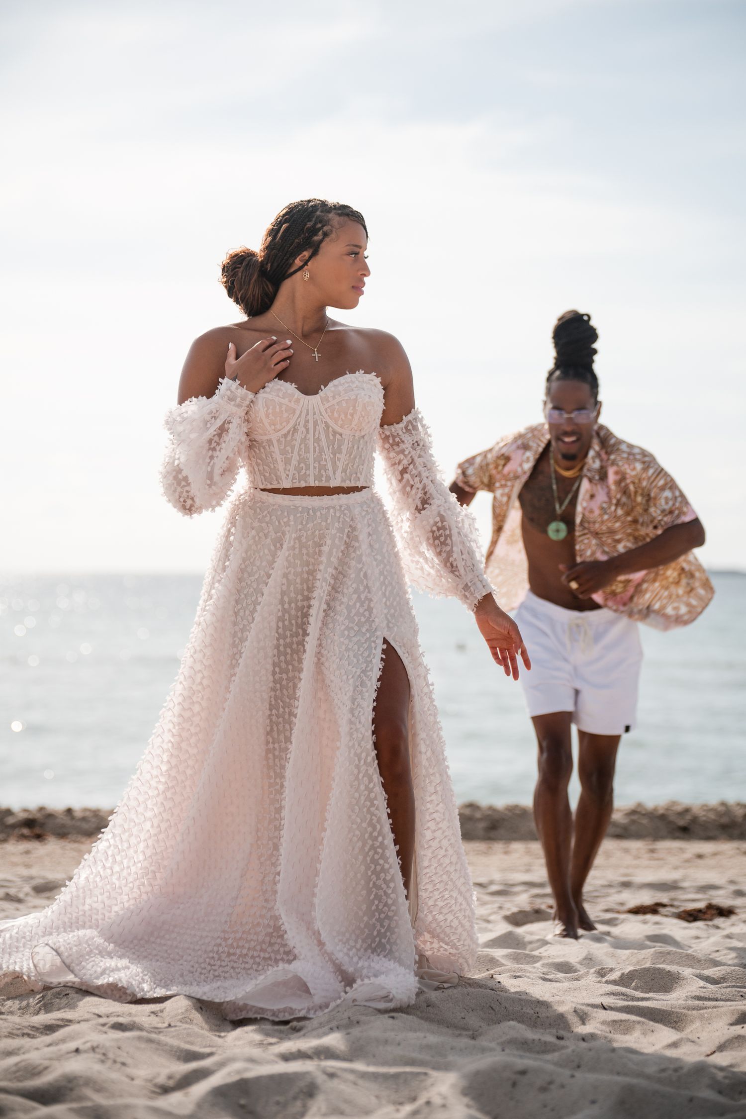 Elegant off-shoulder white lace wedding dress with sheer details photographed on a sunny beach.