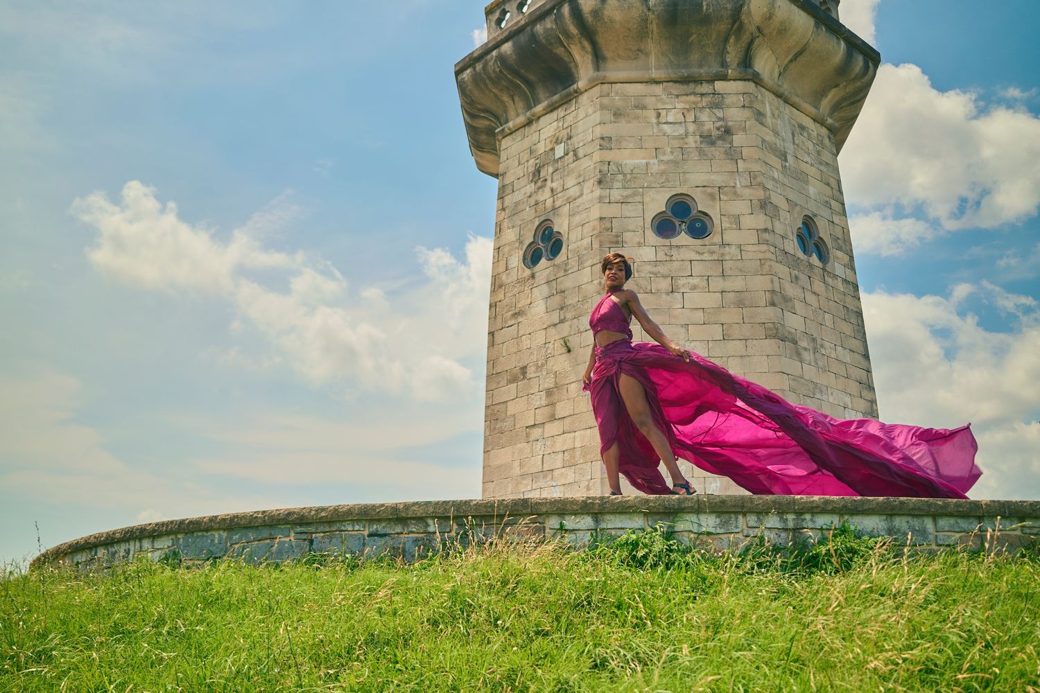 Pink flowing dress billows dramatically against stone tower and green hillside landscape.
