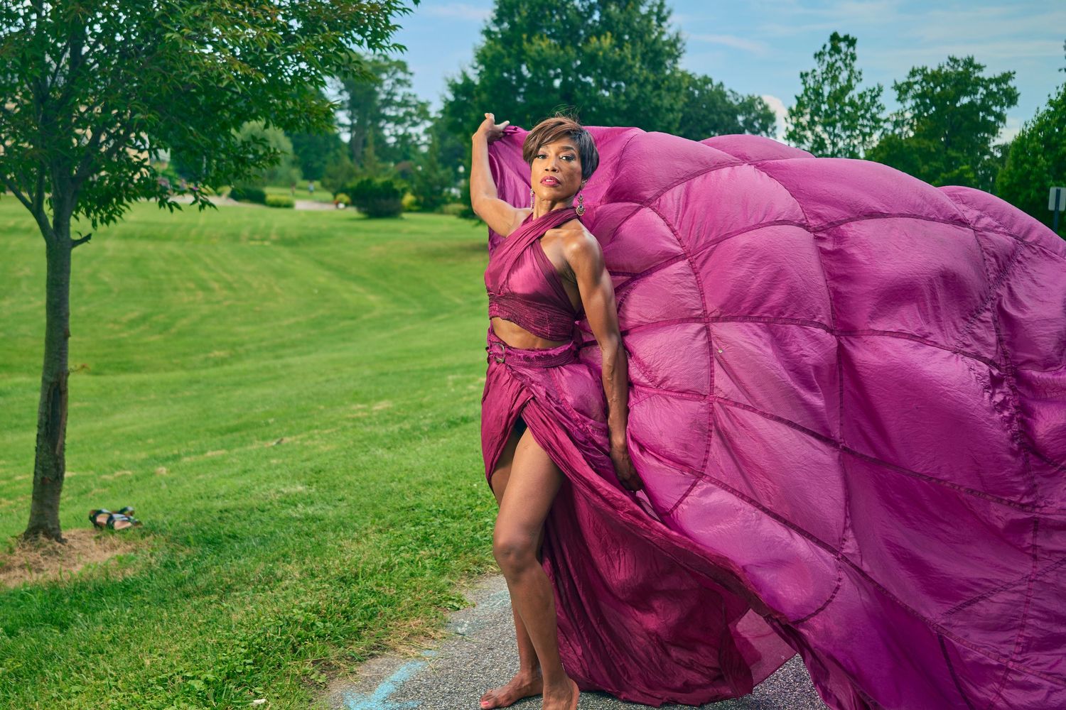 Woman in flowing magenta gown poses outdoors on green lawn with fabric billowing dramatically in the wind.