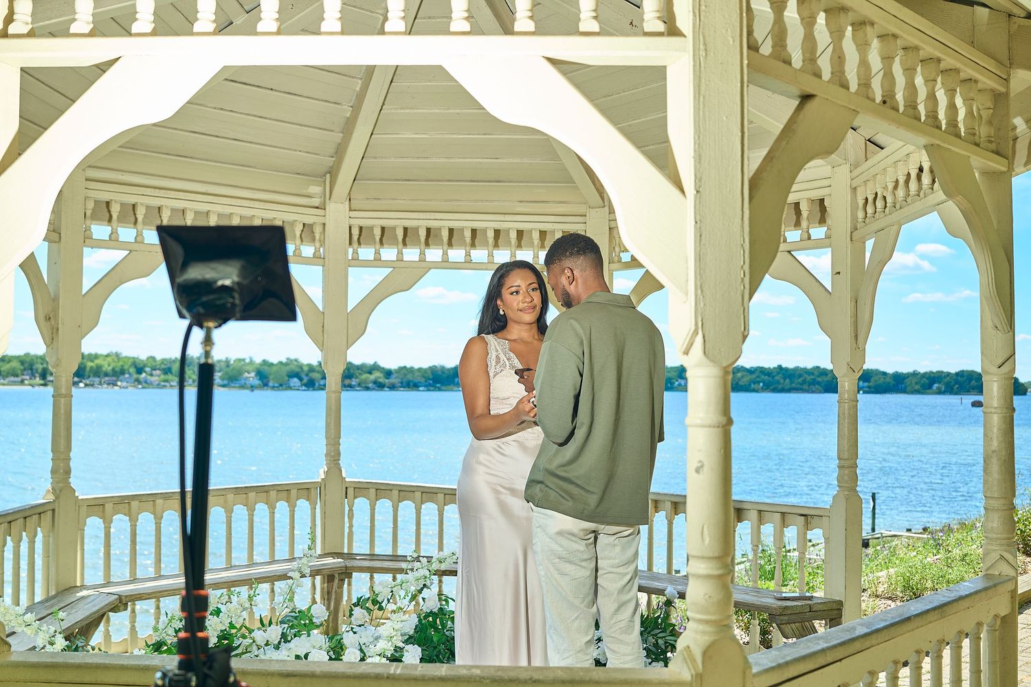 Couple shares a romantic moment in a white gazebo overlooking a serene lake on a sunny day.