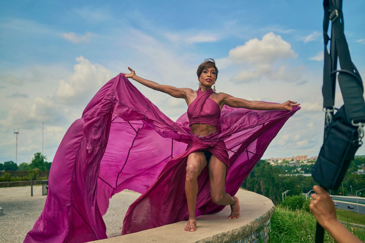 A woman in a flowing magenta dress poses dramatically outdoors, arms spread wide, fabric billowing in the wind.