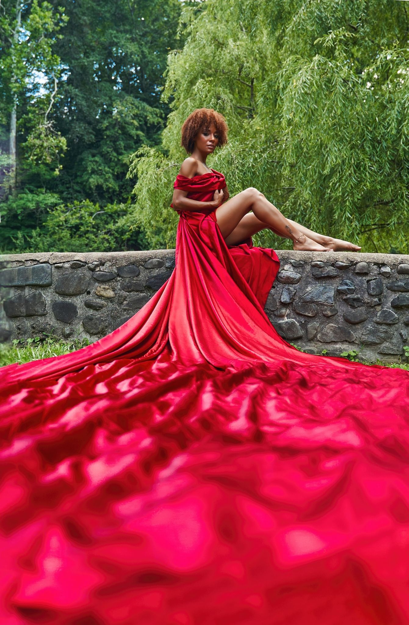 Stunning red satin gown flows dramatically over stone wall beneath willow trees in artistic portrait.