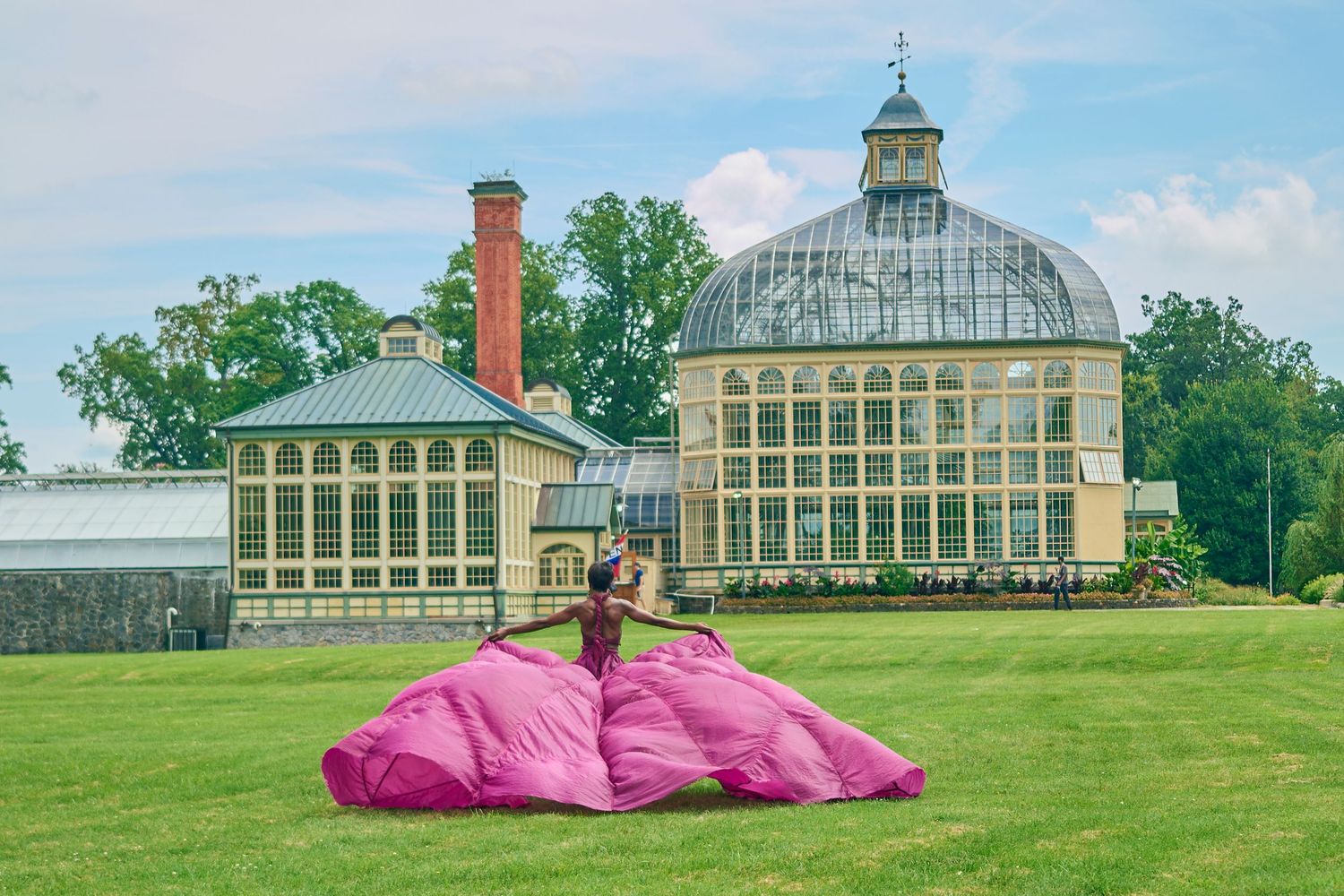 Woman in voluminous pink gown poses on green lawn before elegant domed greenhouse and historic yellow building.