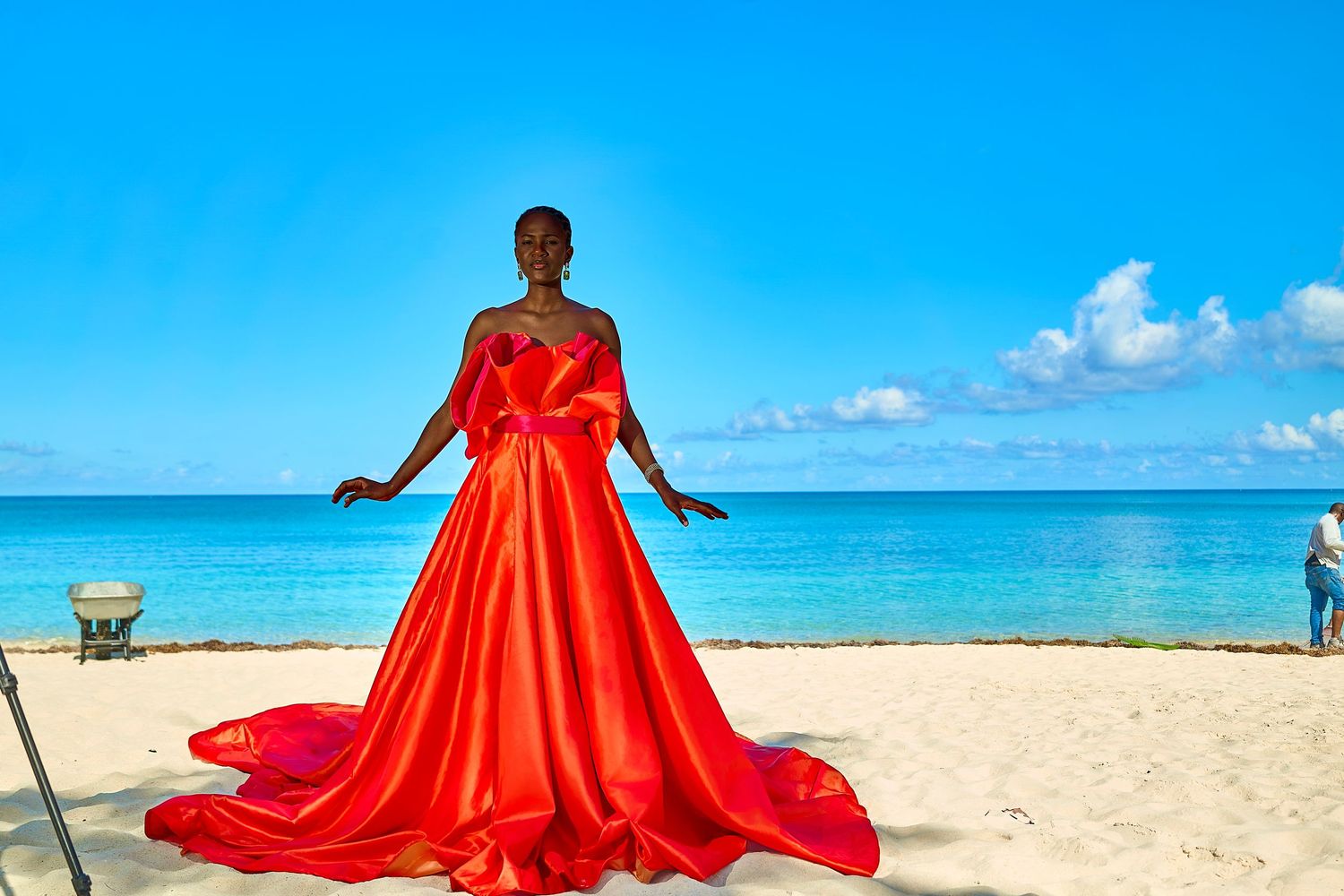 Woman in stunning red ballgown with train poses on sandy beach with turquoise ocean backdrop.