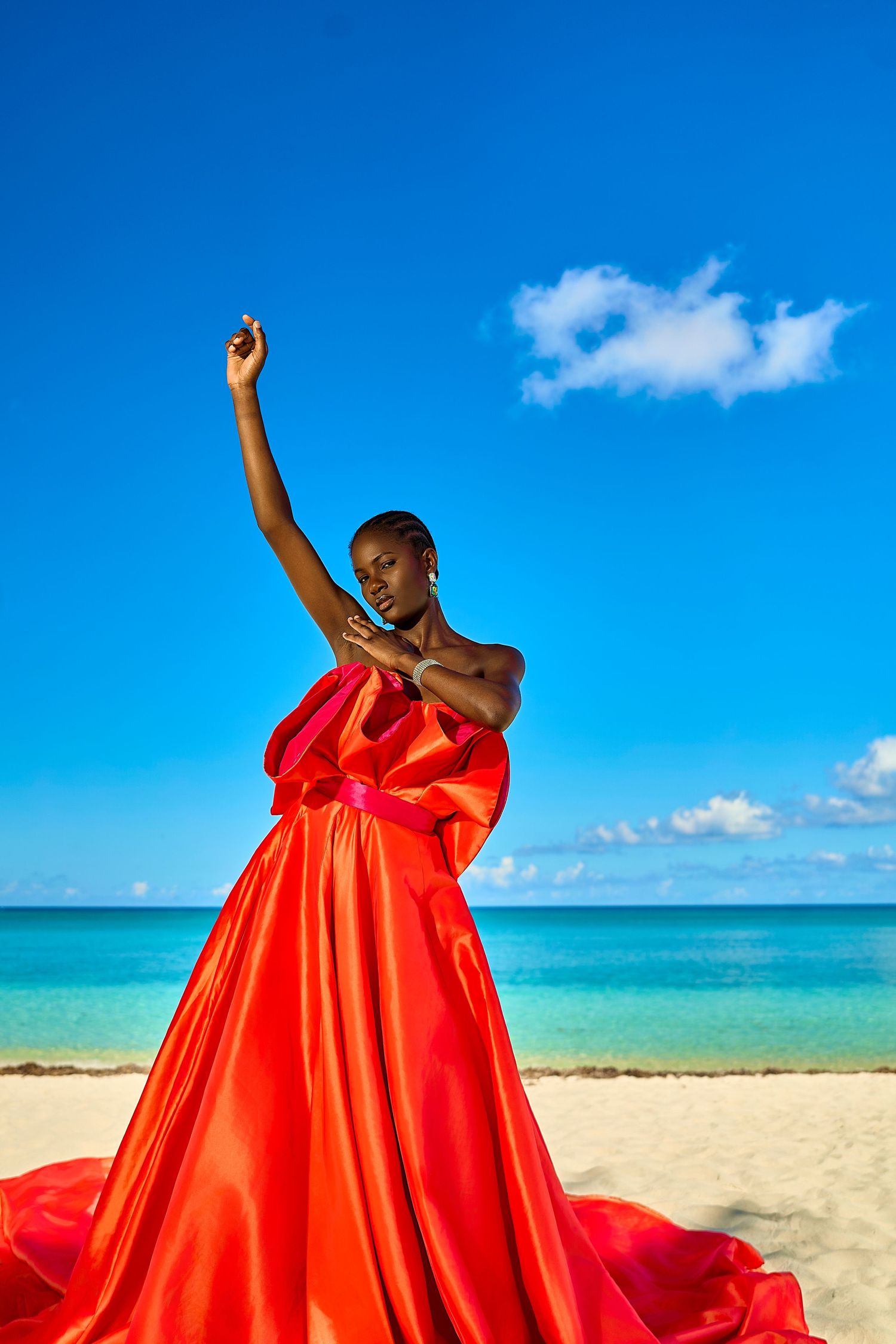A Black woman in a flowing red gown poses gracefully on a tropical beach with turquoise water and blue sky.