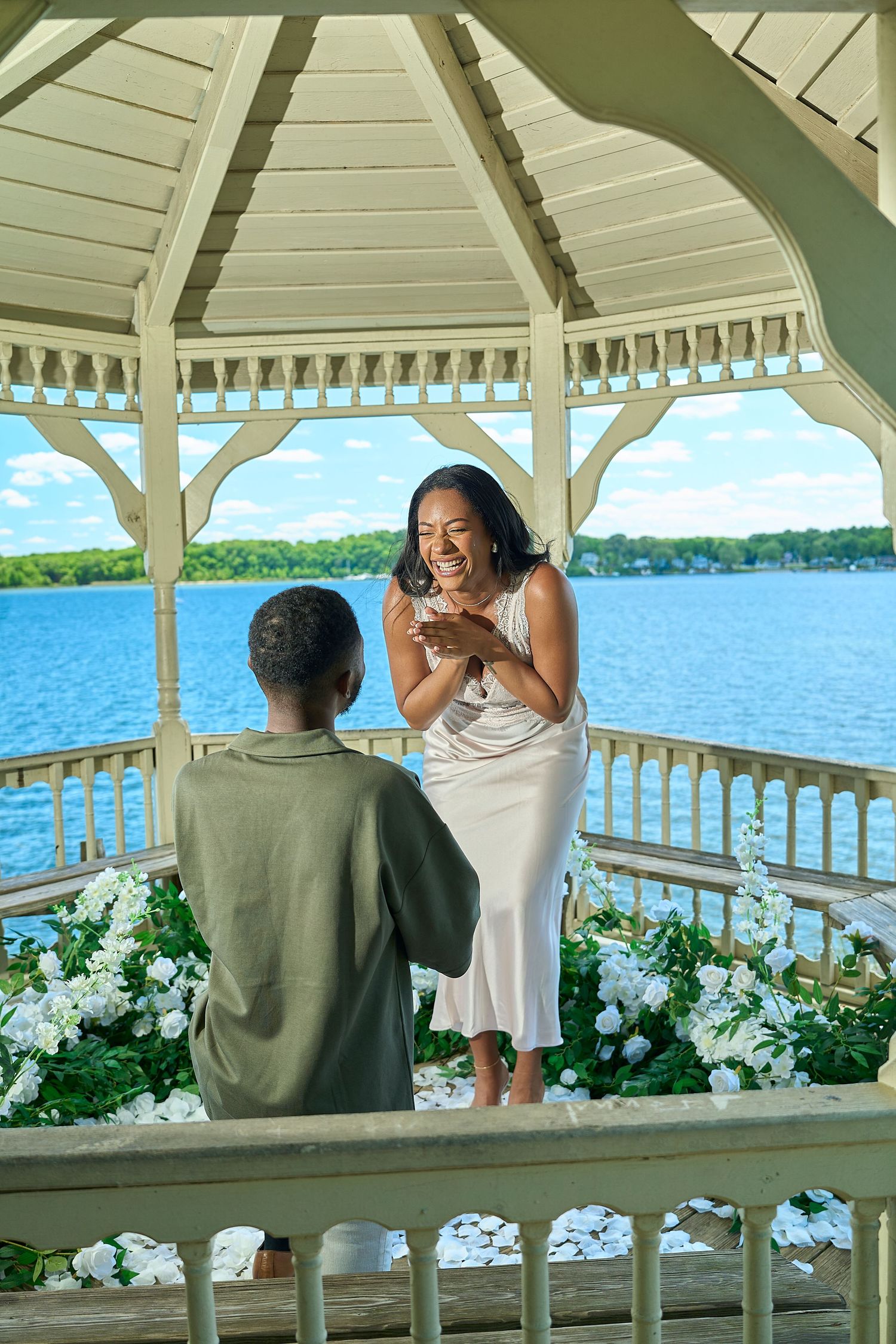 Man kneeling proposing to a joyful woman in a flower-filled lakeside gazebo.