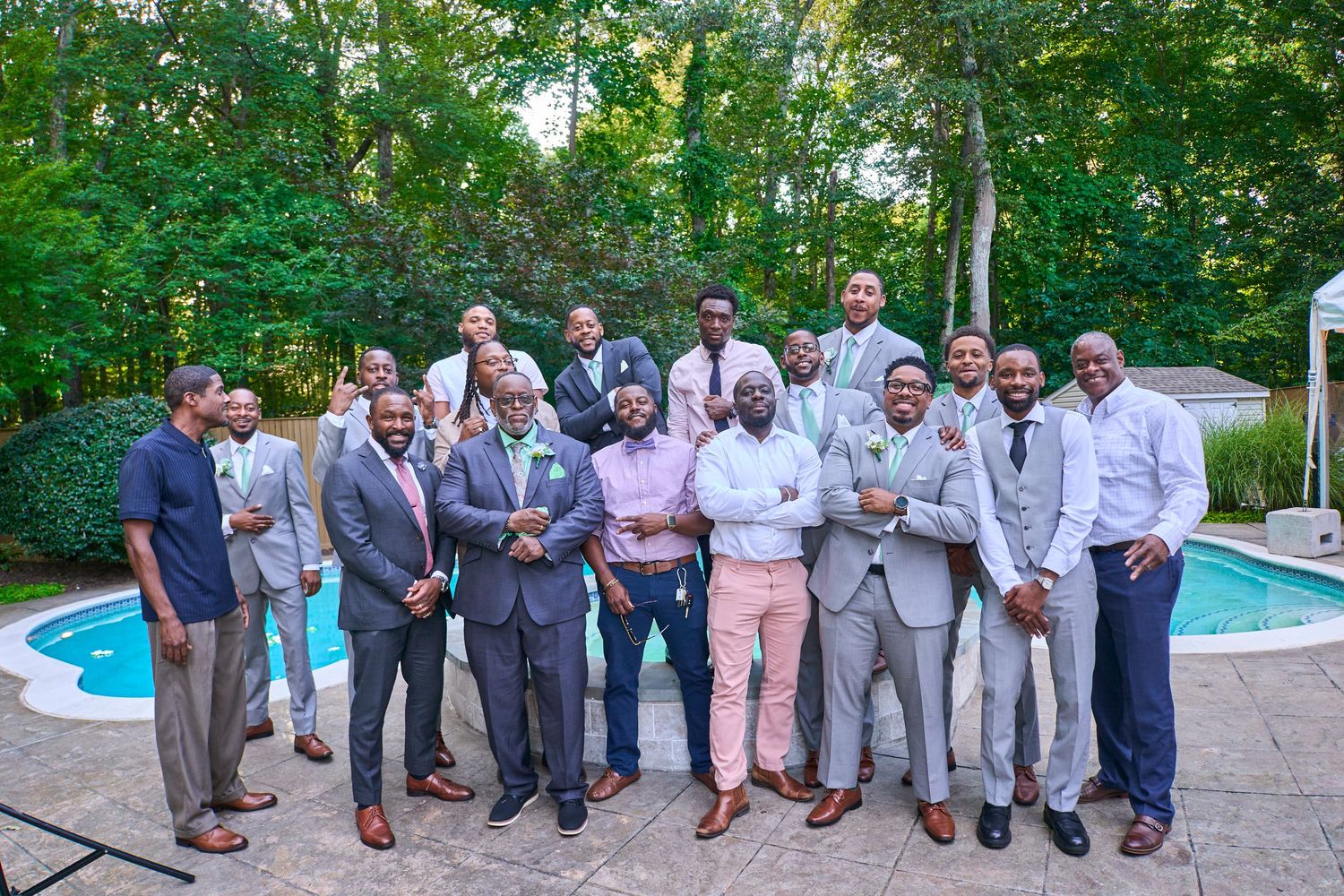 Wedding party in gray suits poses outdoors near a swimming pool surrounded by trees.