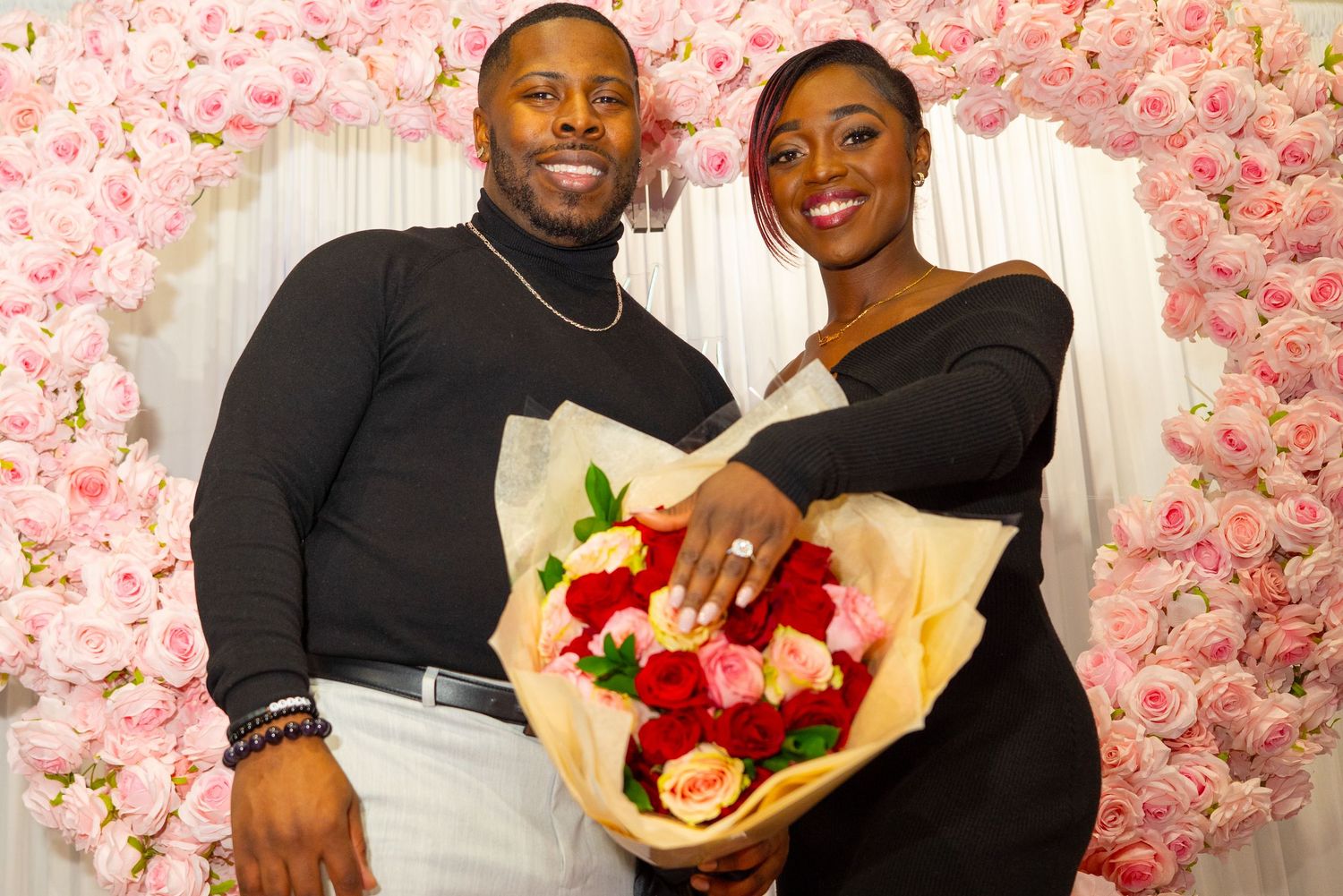 Couple poses with red and pink roses bouquet against floral wall backdrop.