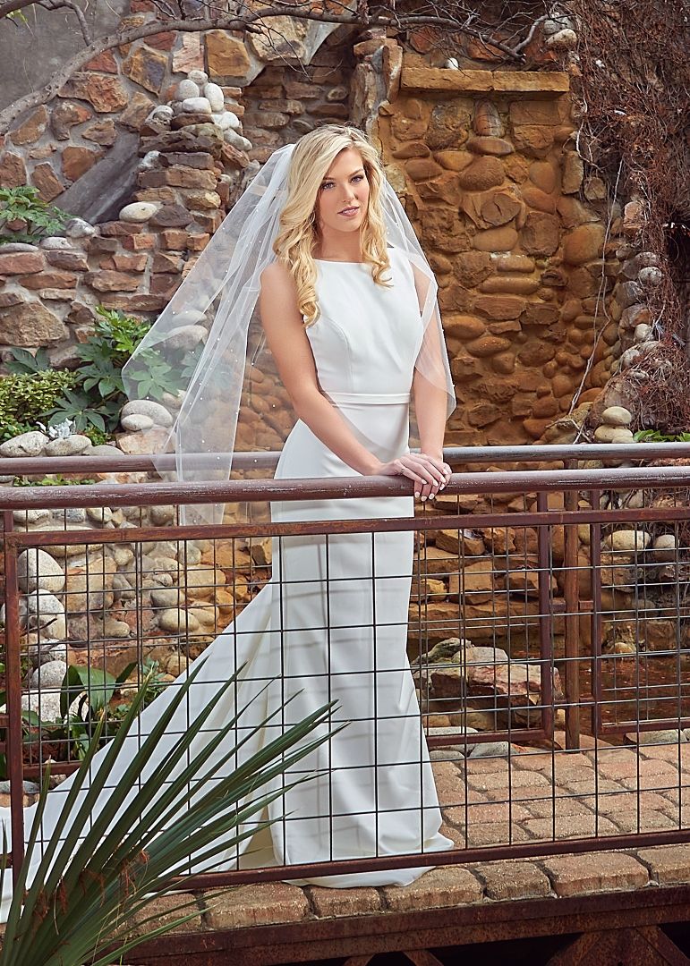 A bride in a white wedding dress poses on a rustic wooden bridge against a stone wall with palm plants in view.