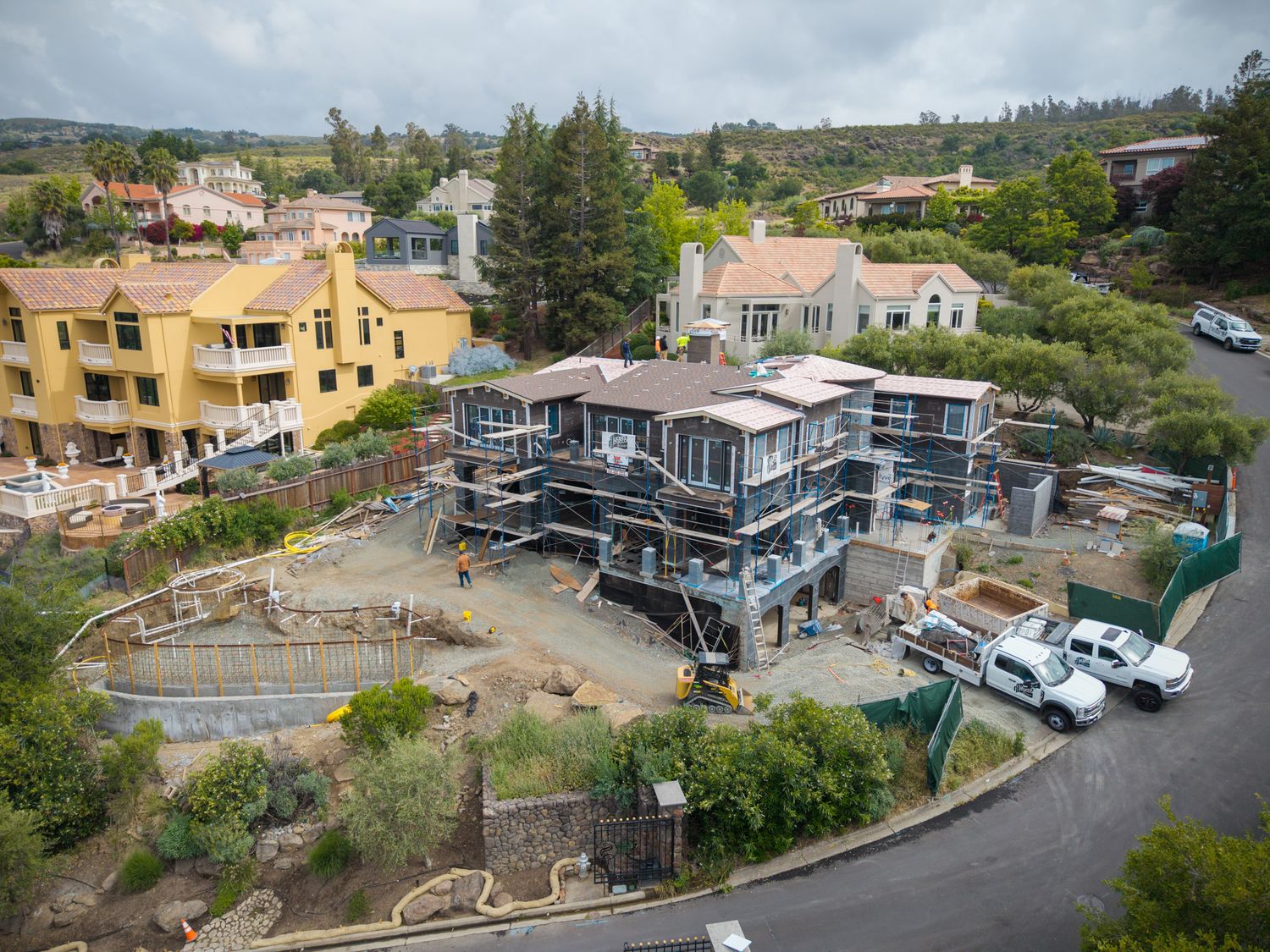 Aerial view of a multi-story home under construction in a residential hillside neighborhood with scaffolding and parked vehicles.