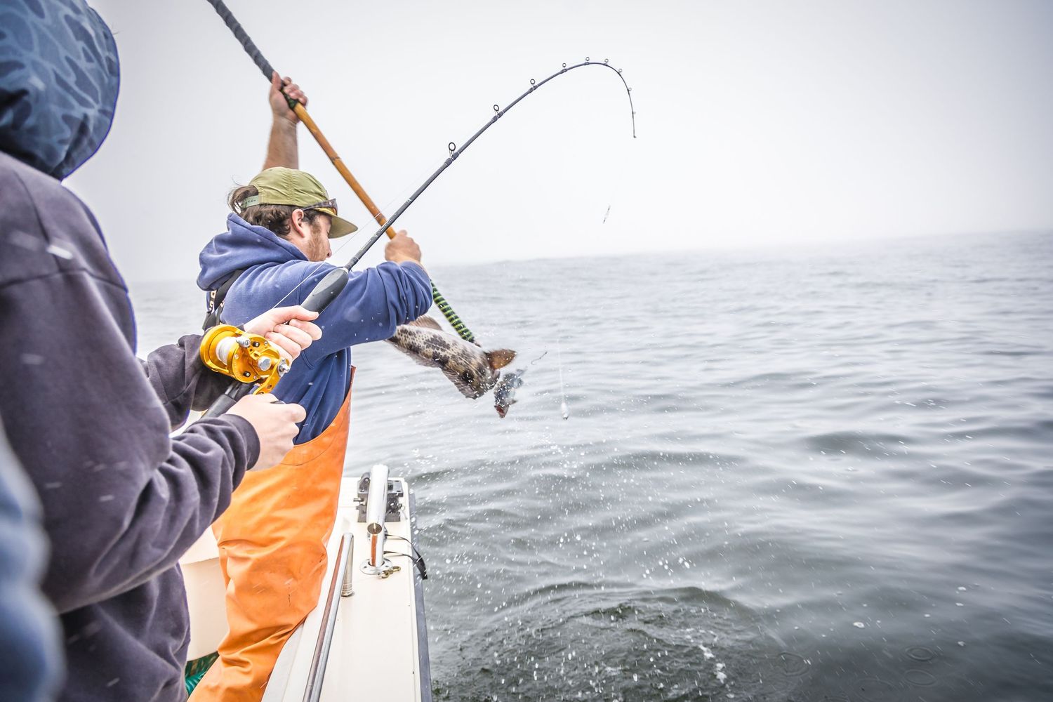 Deep sea fisherman in orange bibs leans over boat railing with fishing rod during overcast day at sea.
