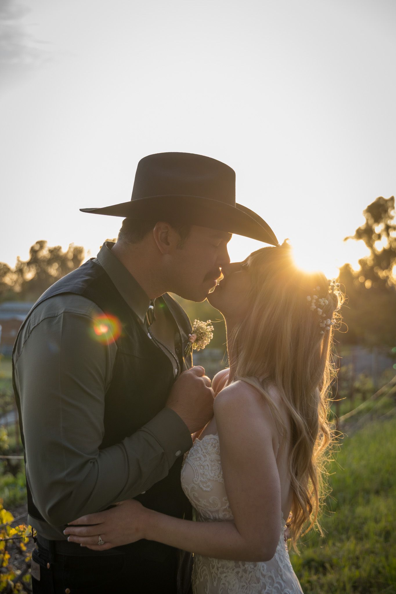Golden sunset silhouettes a romantic couple embracing outdoors, featuring cowboy hat and white dress.