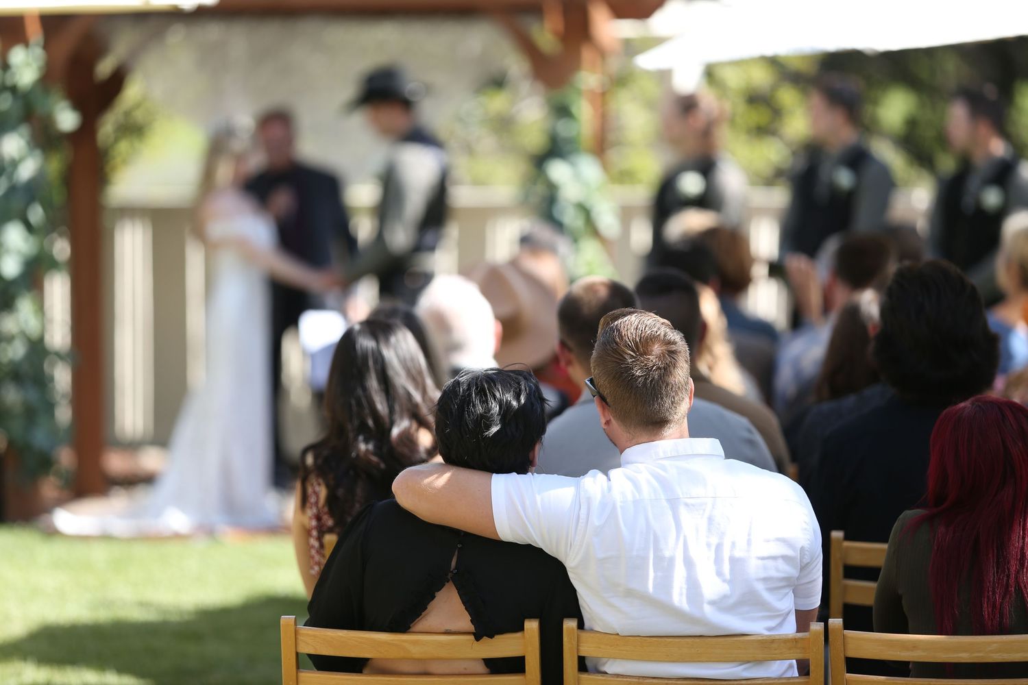 Wedding guests sitting on wooden chairs at an outdoor garden ceremony during a sunny afternoon.