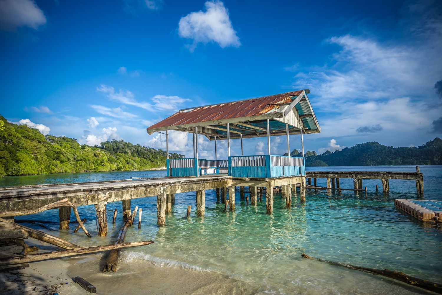 Weathered wooden pier stretching across crystal clear turquoise waters with mountains in background.