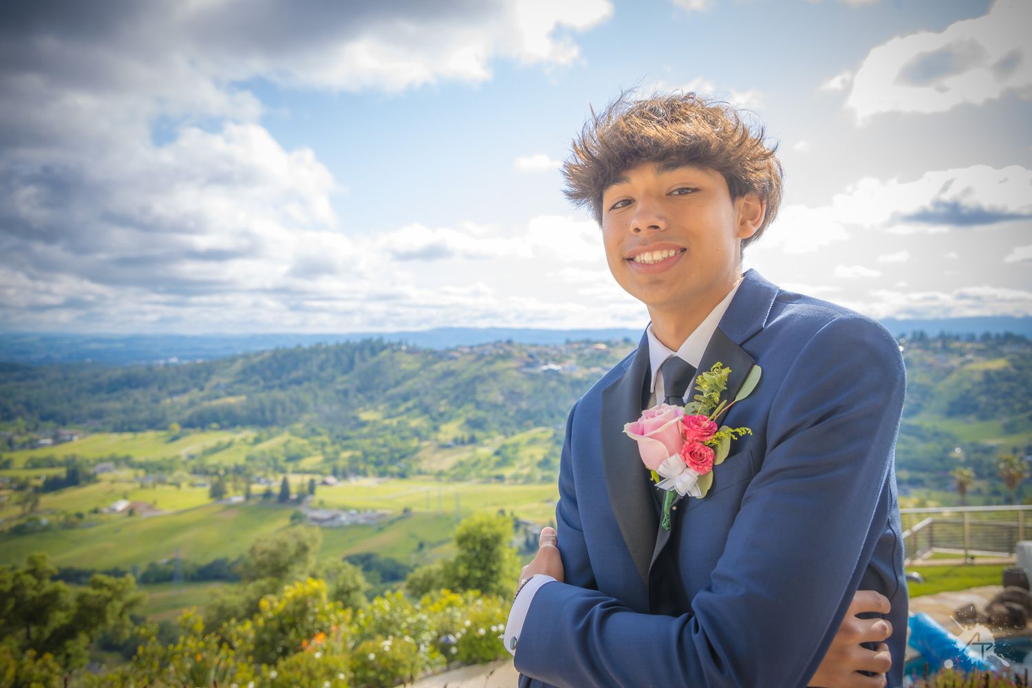 Person in navy suit with pink boutonniere smiles against mountainous backdrop.