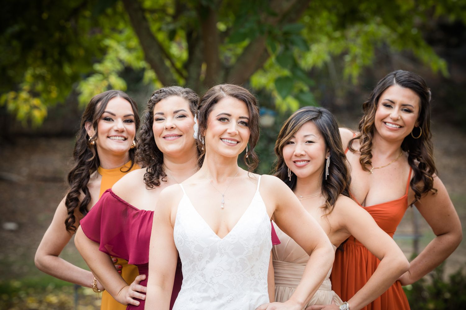 A group of bridesmaids in burgundy and rust colored dresses pose with a bride in a white lace gown outdoors.