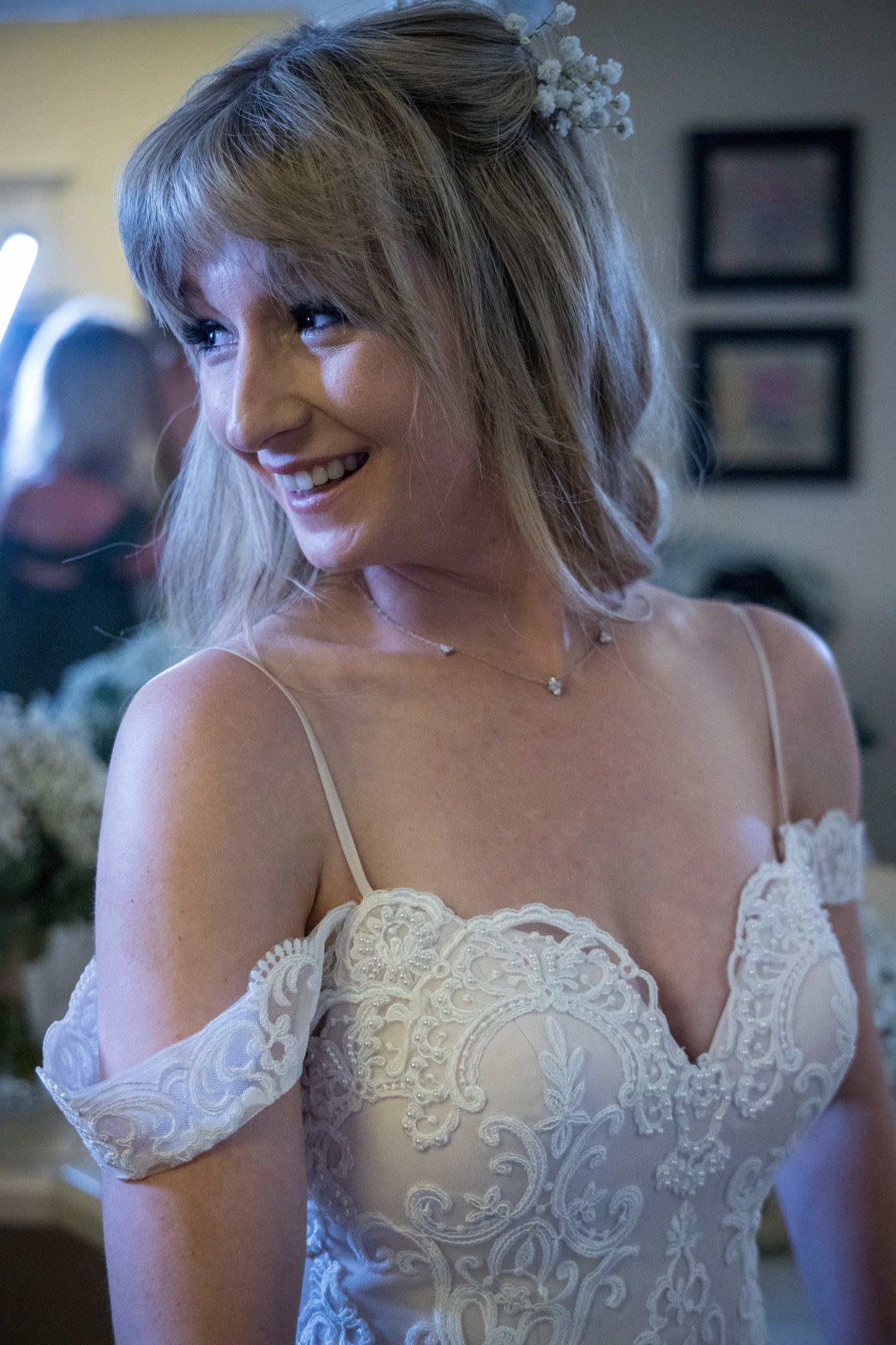 Bride wearing delicate off-shoulder white lace wedding dress smiles warmly in natural lighting.