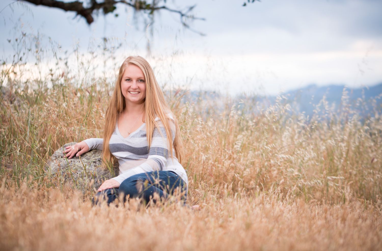 Person sitting in a golden field of tall grass with mountains visible in the background on a sunny day.