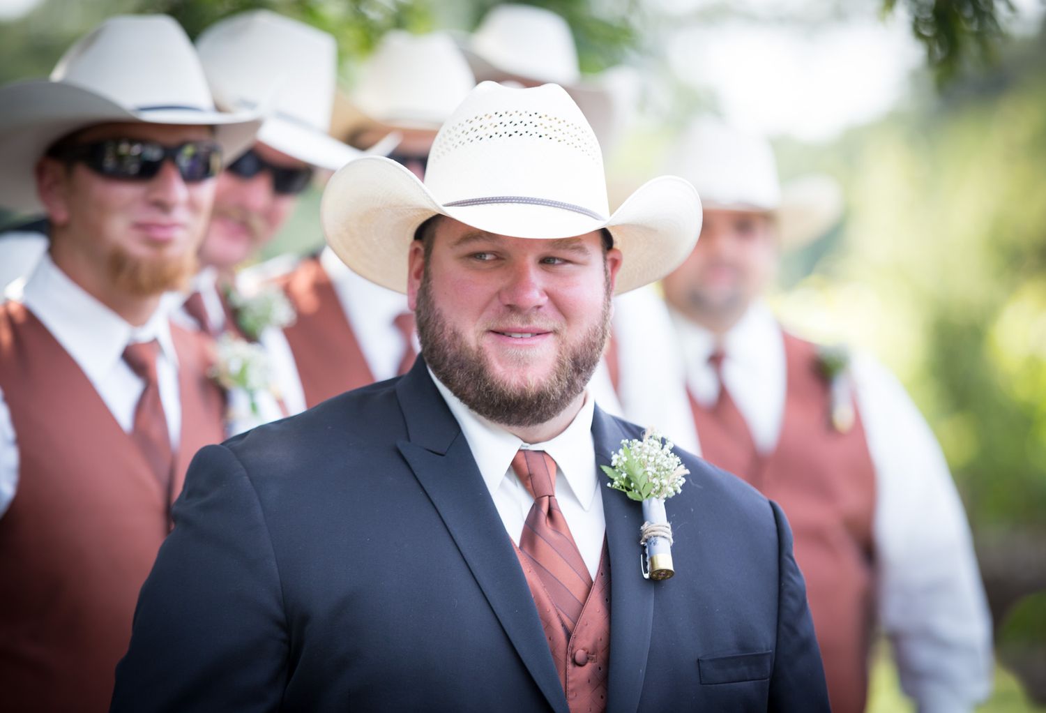 A groom in a black suit and rust-colored tie poses with groomsmen wearing cowboy hats at an outdoor wedding.