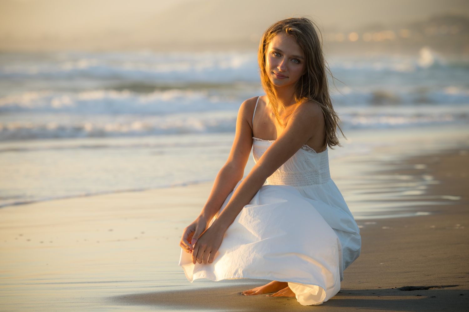 Young woman in white dress crouching on sandy beach at golden sunset with ocean waves in background.