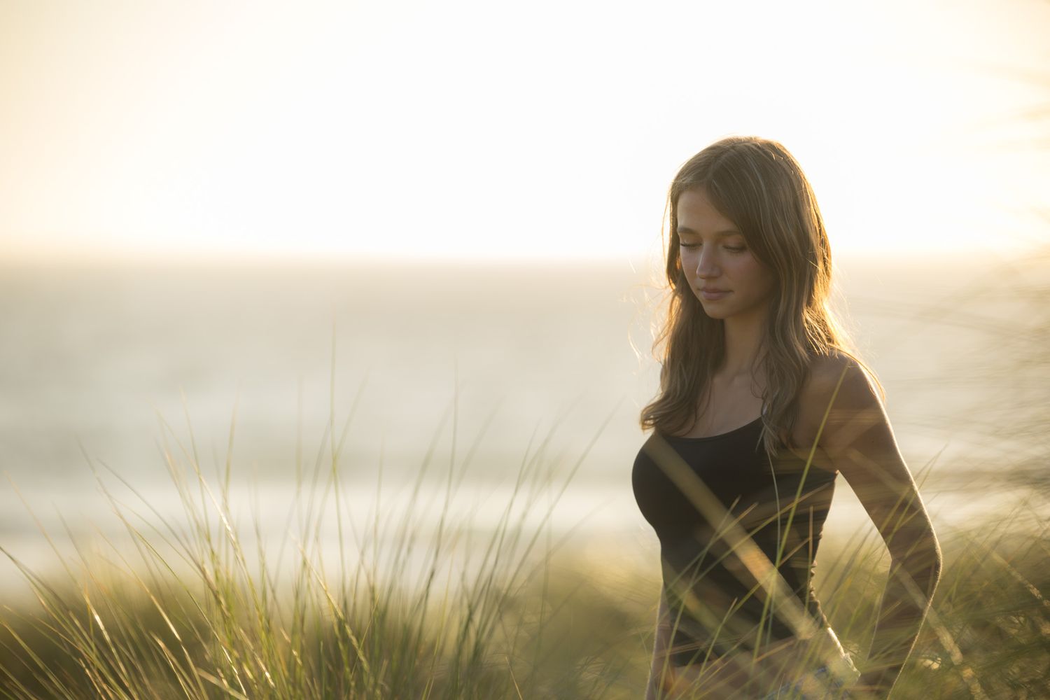 Young woman with long brown hair standing in golden sunset light among tall grass near the ocean.