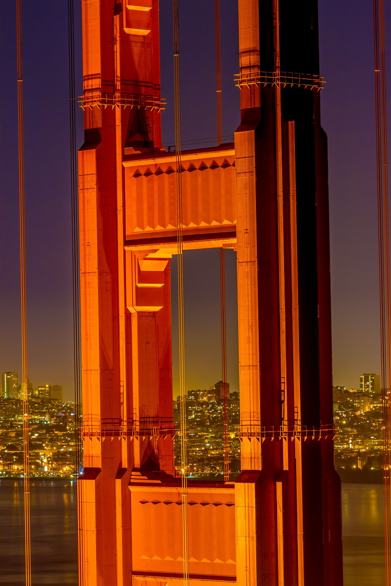 Golden Gate Bridge tower detail illuminated in warm orange light with city lights twinkling behind.