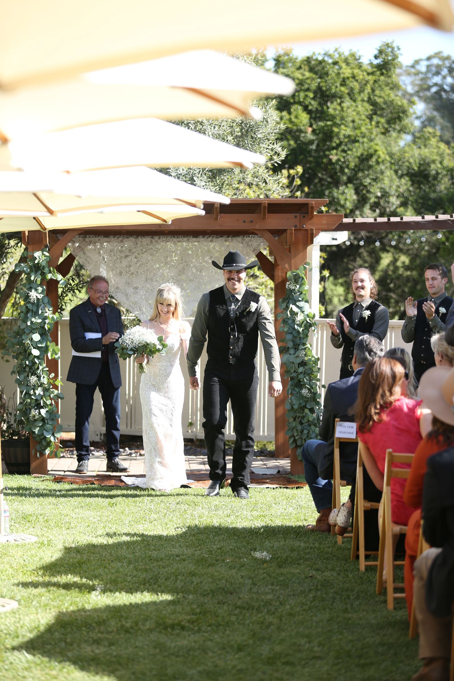 An outdoor wedding ceremony under a wooden pergola with white umbrellas and floral decorations on a green lawn.