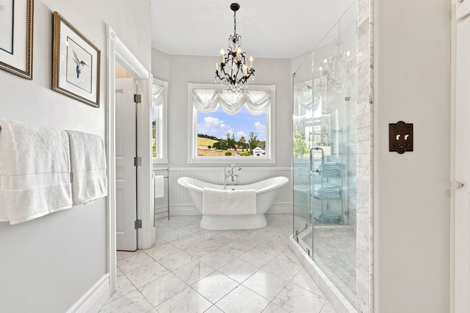 Elegant white bathroom with freestanding soaking tub, glass shower, chandelier, marble tile floors, and a bright window.