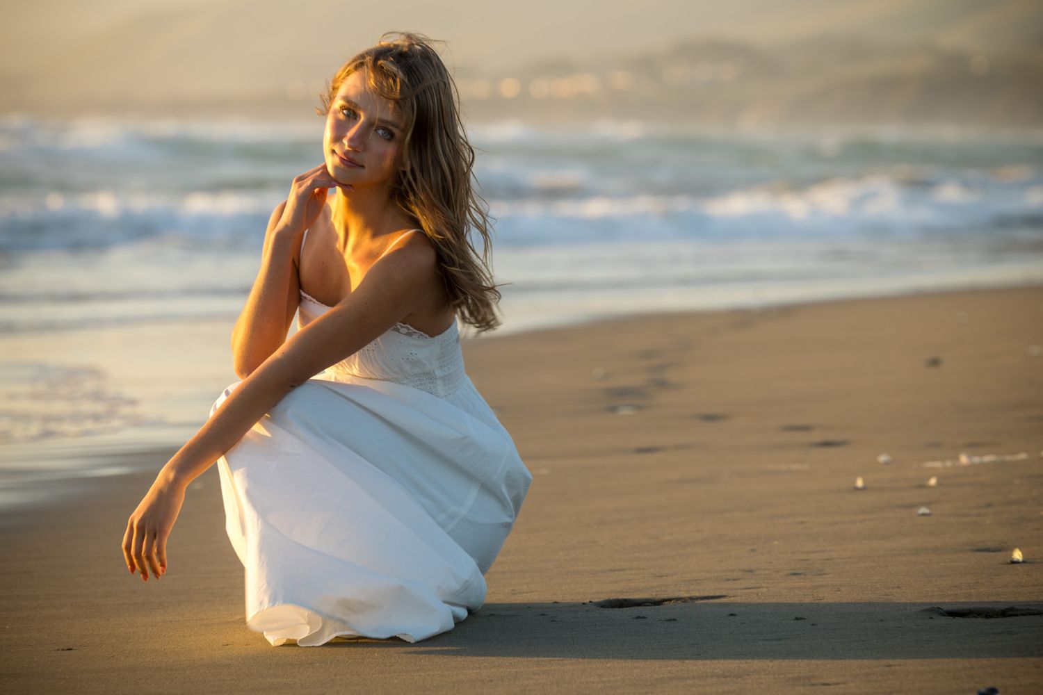 A woman in a white dress crouches on a sandy beach at golden hour with ocean waves in the background.