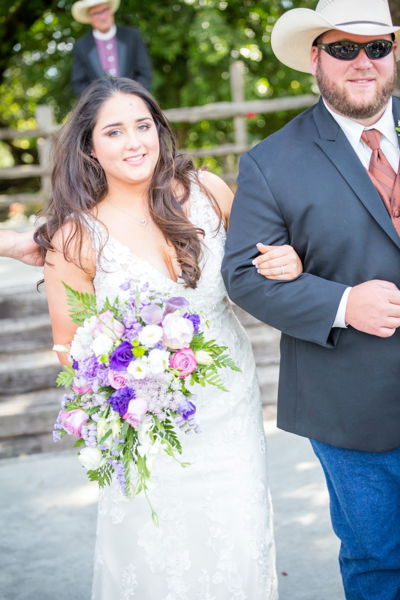 A couple in western wedding attire stands with a vibrant purple and pink floral bouquet.